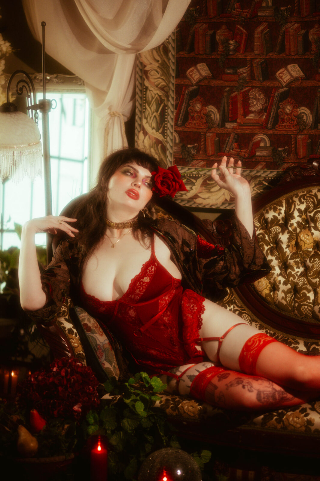 A model reclines in a vintage armchair wearing a red lace lingerie set with thigh high stockings, a rose tucked beside her face. Soft window light and library inspired decor highlight one of our favorite boudoir session tips for posing with props. Royal Lune Photo.