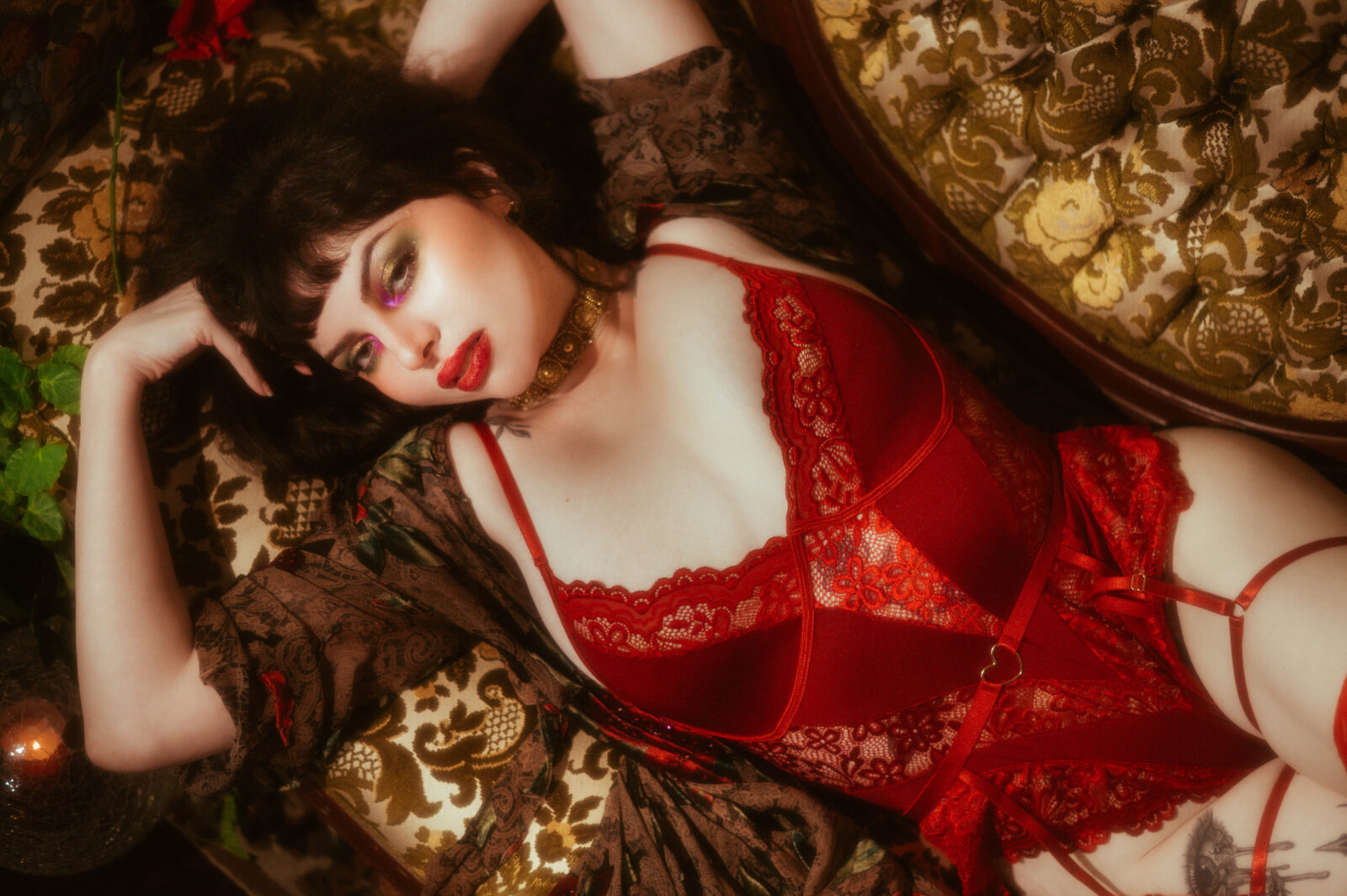 A close up portrait shows a model lying back in red lace lingerie with a gold collar necklace and bold red lip against a patterned vintage backdrop. This image illustrates the kind of glamorous styling covered in our boudoir prep guide. Royal Lune Photo.