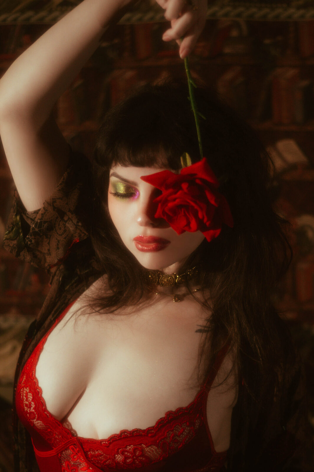 A close up beauty shot features a model with dark hair and dramatic eye makeup holding a red rose over one eye while wearing red lace lingerie. Soft golden lighting adds to the romantic mood featured throughout this boudoir prep guide. Royal Lune Photo.
