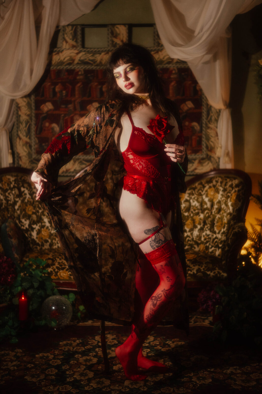 A full length portrait captures a model in red lace lingerie and a sheer floral robe standing in front of a vintage sofa holding a red rose. This confident standing pose is a wonderful reference as you prepare for your boudoir session. Royal Lune Photo.