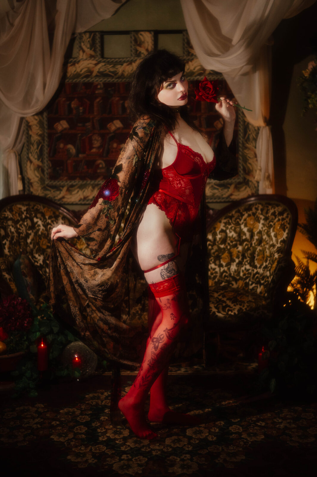 A model wearing a red lace bodysuit and sheer printed robe stands beside an antique tufted sofa holding a red rose. Candlelight and rich tapestry decor set the scene for this guide to help you prepare for your boudoir session. Royal Lune Photo.