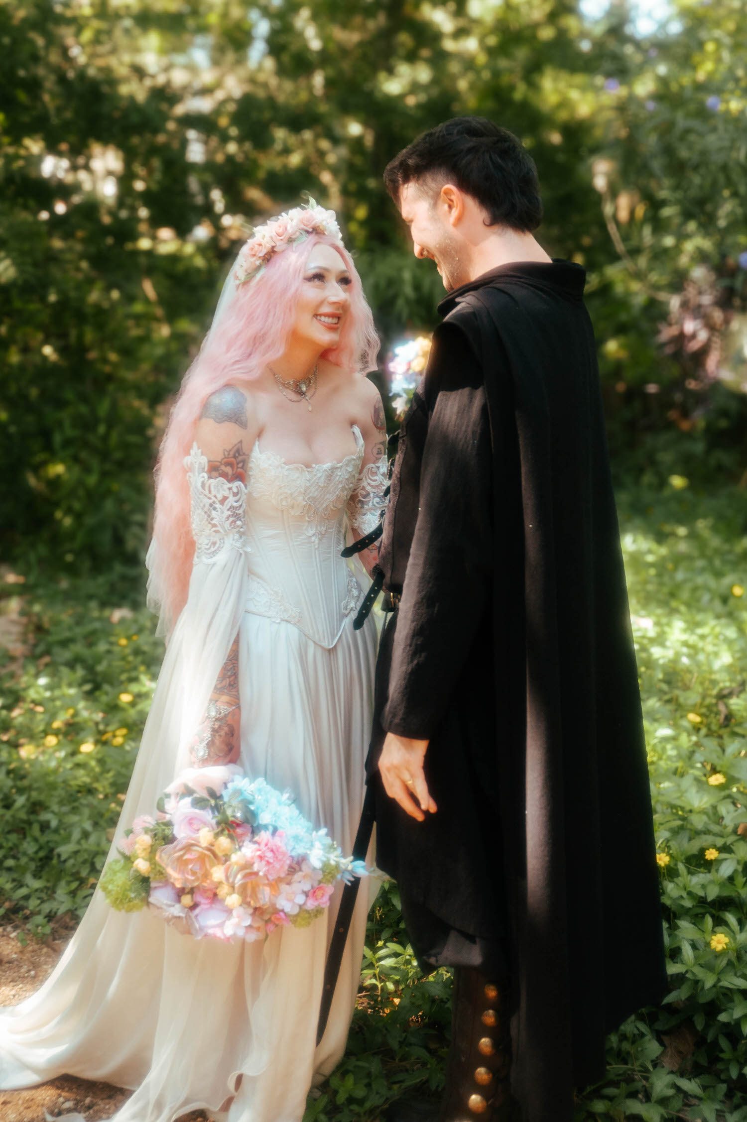 The bride and groom share a laugh in a sunlit wooded area, her pink hair and floral crown contrasting beautifully with his black cloak and medieval style jacket. Portraits like this are a favorite part of any Texas Renaissance Festival wedding. Royal Lune Photo.