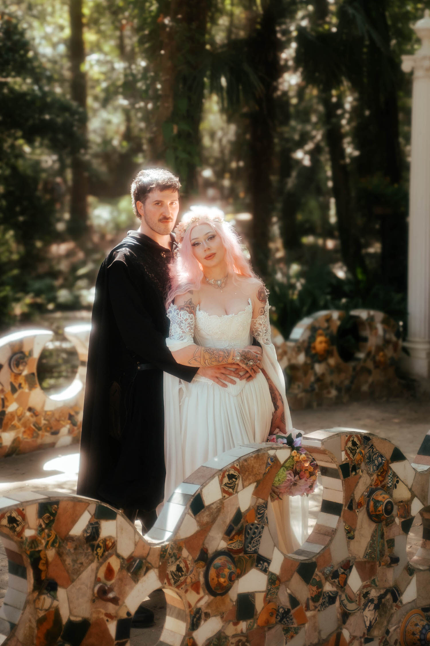 The newlyweds pose together by the colorful mosaic bench, the groom in his black medieval coat and the bride in her Firefly Path Bridal Gown with her bouquet resting nearby. This whimsical garden setting is a hidden gem for a Texas Renaissance Festival wedding. Royal Lune Photo.