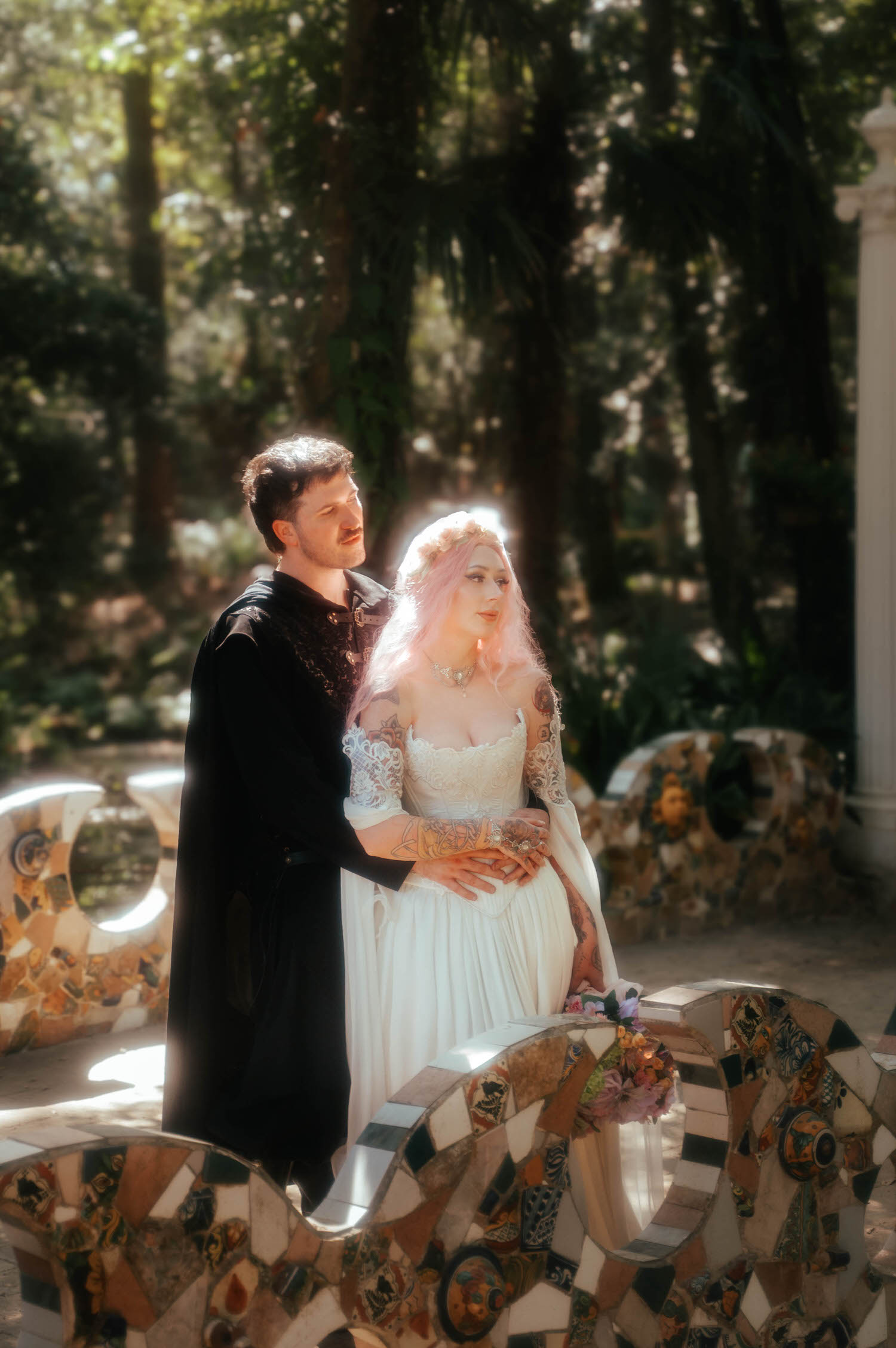 The bride and groom stand together near a mosaic tiled bench in a sunlit forest clearing, her tattooed arms and pink hair catching the dappled light. Settings like this showcase why couples choose a Renaissance festival wedding for their big day. Royal Lune Photo.