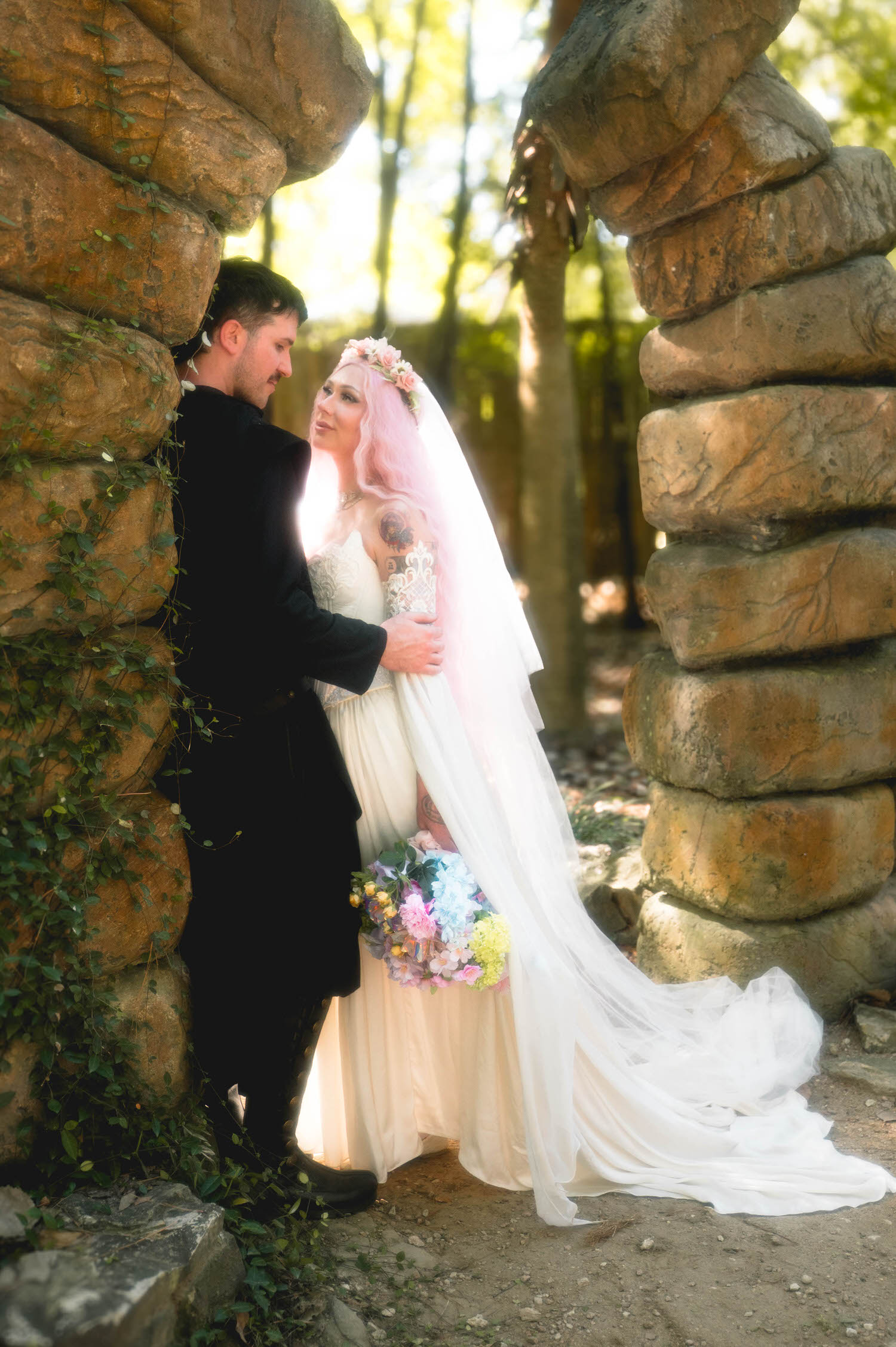 The bride and groom stand framed by a rustic stone archway in the woods, her veil cascading down and her bouquet of pastel flowers in hand. Dramatic natural backdrops like this are part of what makes a Texas Renaissance Festival wedding photographer's portfolio so striking. Royal Lune Photo.