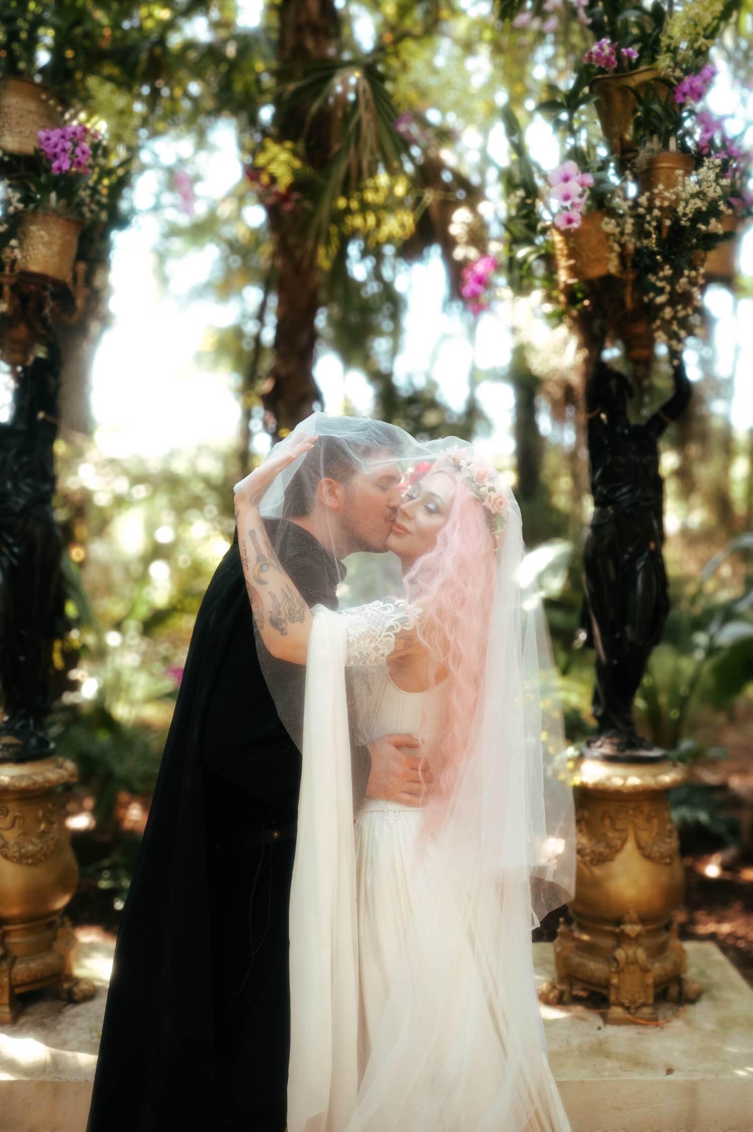 The bride and groom share a kiss under her veil in a wooded garden lined with golden statues and hanging orchids. Romantic details like this are a highlight of any Texas Renaissance Festival wedding photographer's work. Royal Lune Photo.