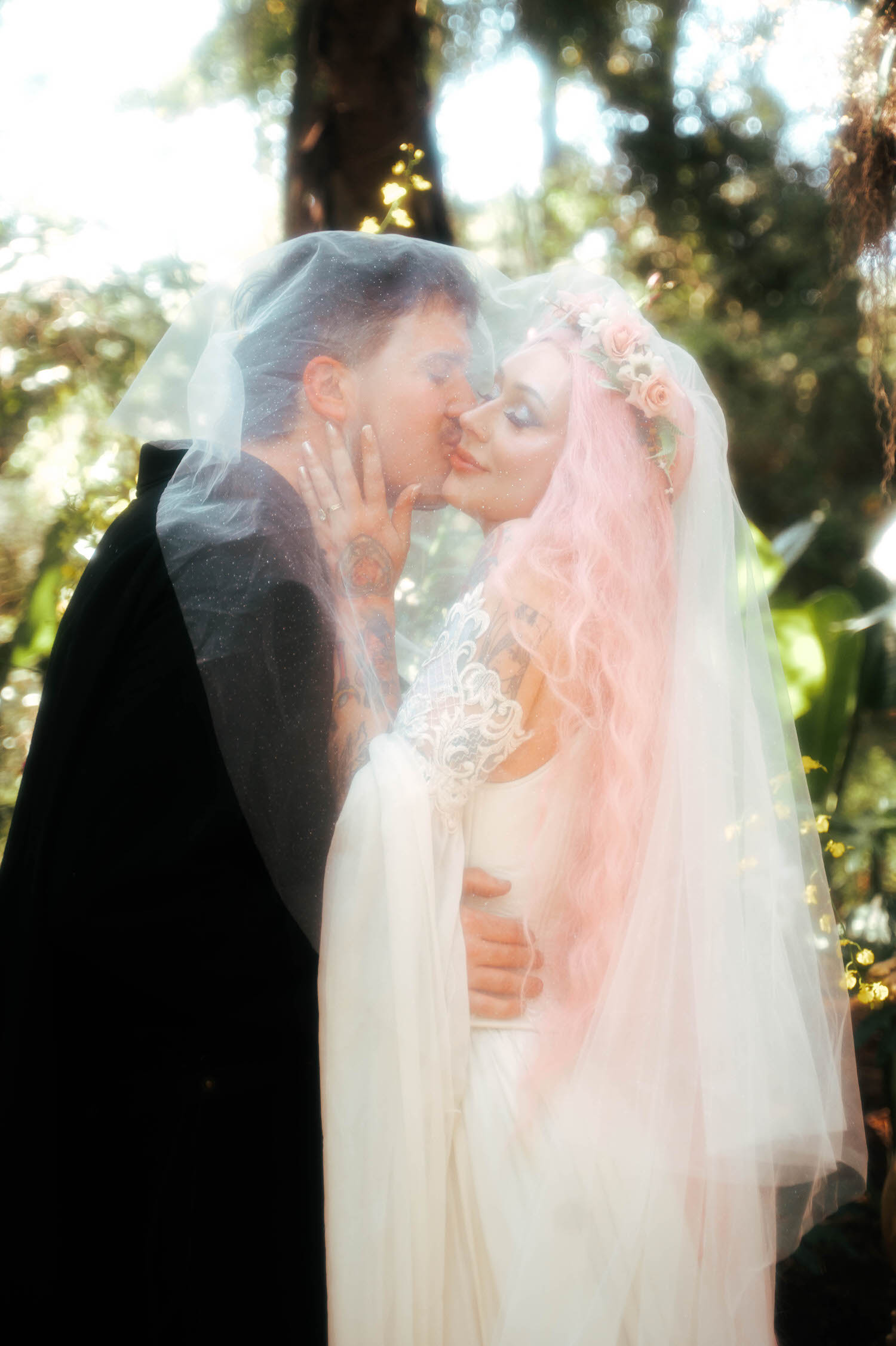 The newlyweds steal a kiss beneath the bride's veil, her pink hair and lace sleeves glowing in the dappled forest light. Intimate moments like this define the artistry of a fantasy wedding photographer. Royal Lune Photo.