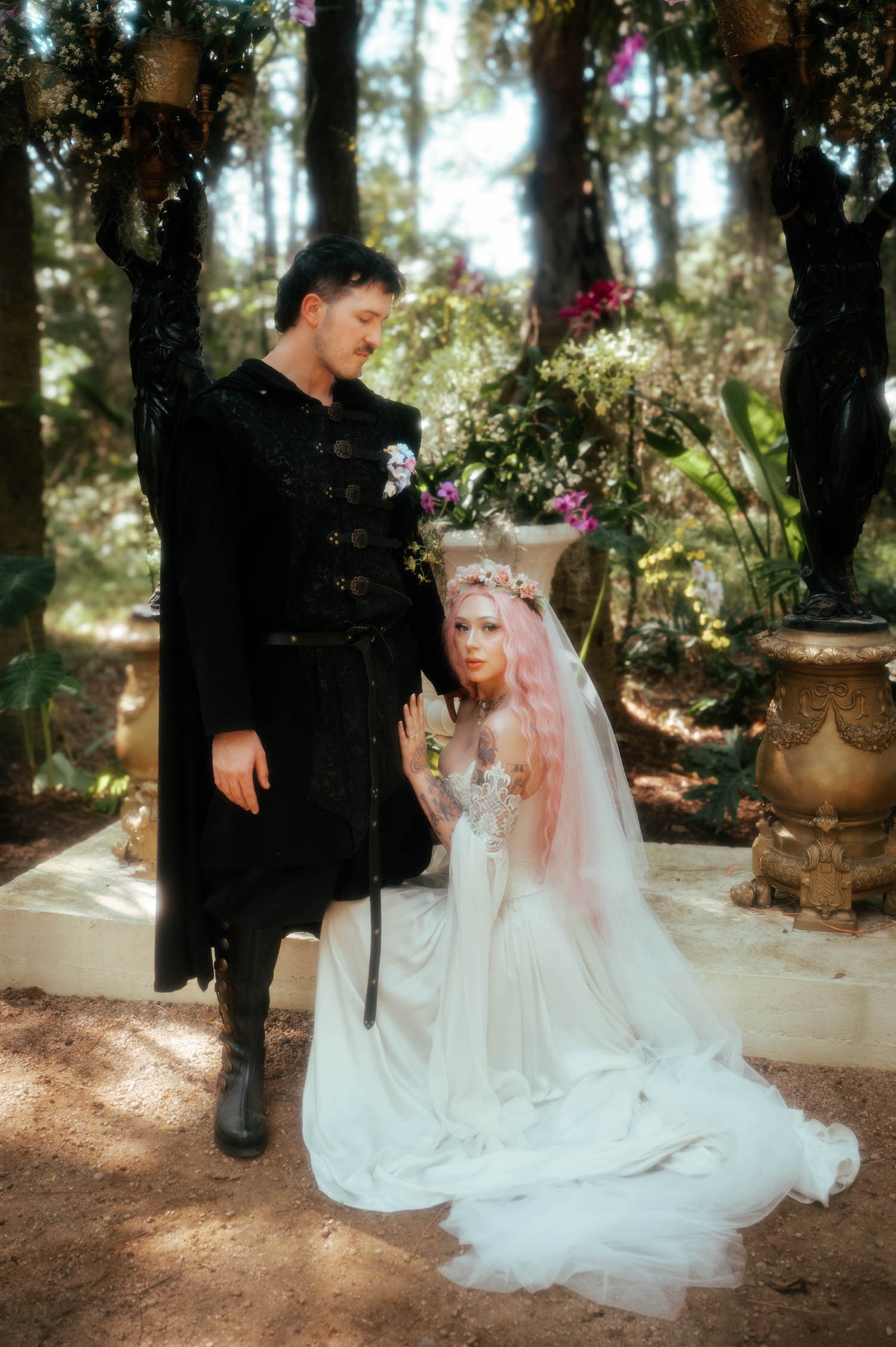 The groom rests his hand on the bride's shoulder as she kneels beside an ornate garden urn filled with orchids, her gown pooling around her on the forest floor. Scenes like this capture the romantic, painterly quality couples look for in a Renaissance festival wedding. Royal Lune Photo.