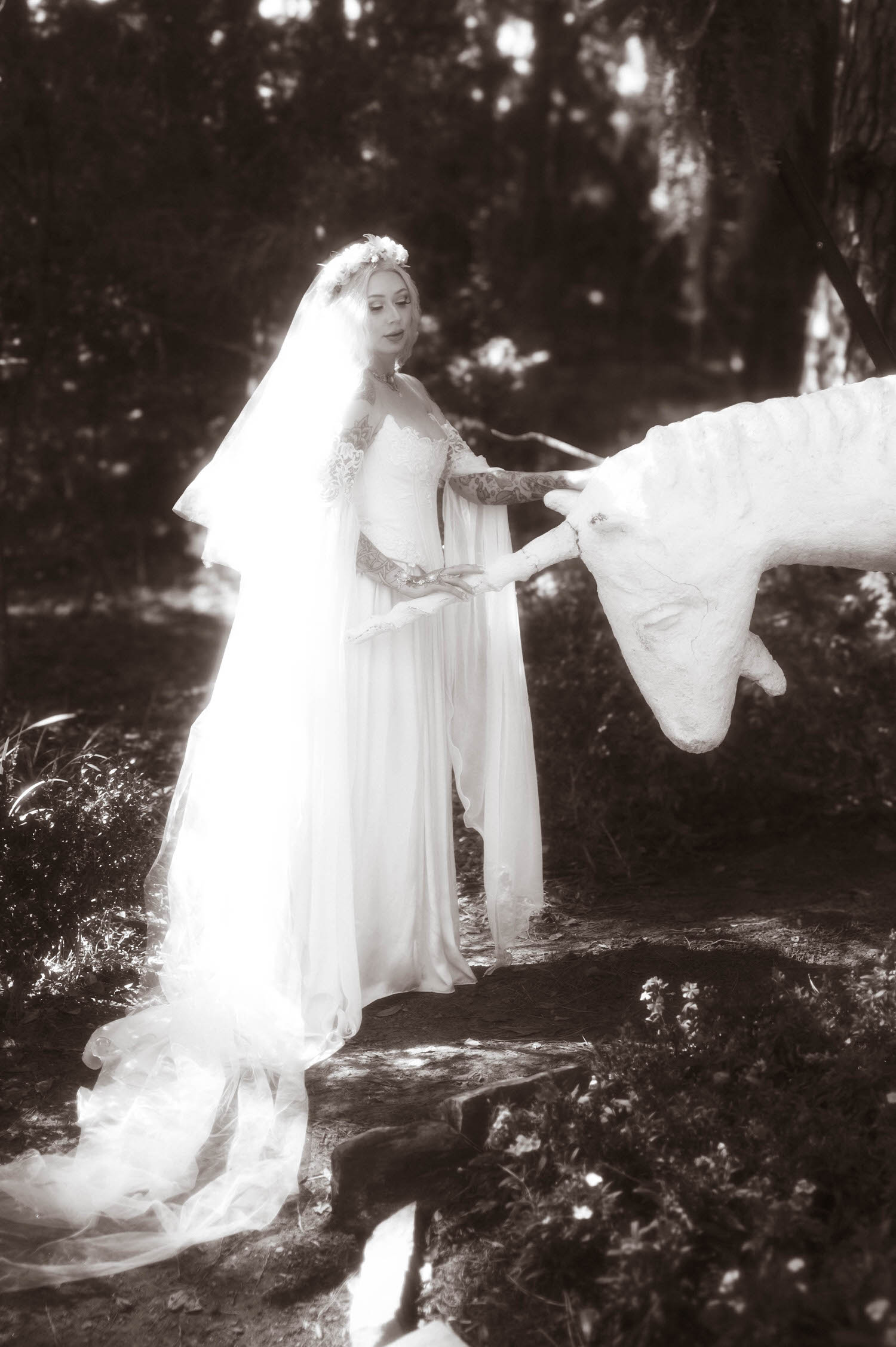 In this black and white portrait, the bride is wearing a Firefly Path bridal gown while gently reaching for the unicorn statue's face, her flower crown and flowing gown adding to the storybook atmosphere. Enchanting moments like this are part of what sets a Texas Renaissance Festival wedding apart. Royal Lune Photo.