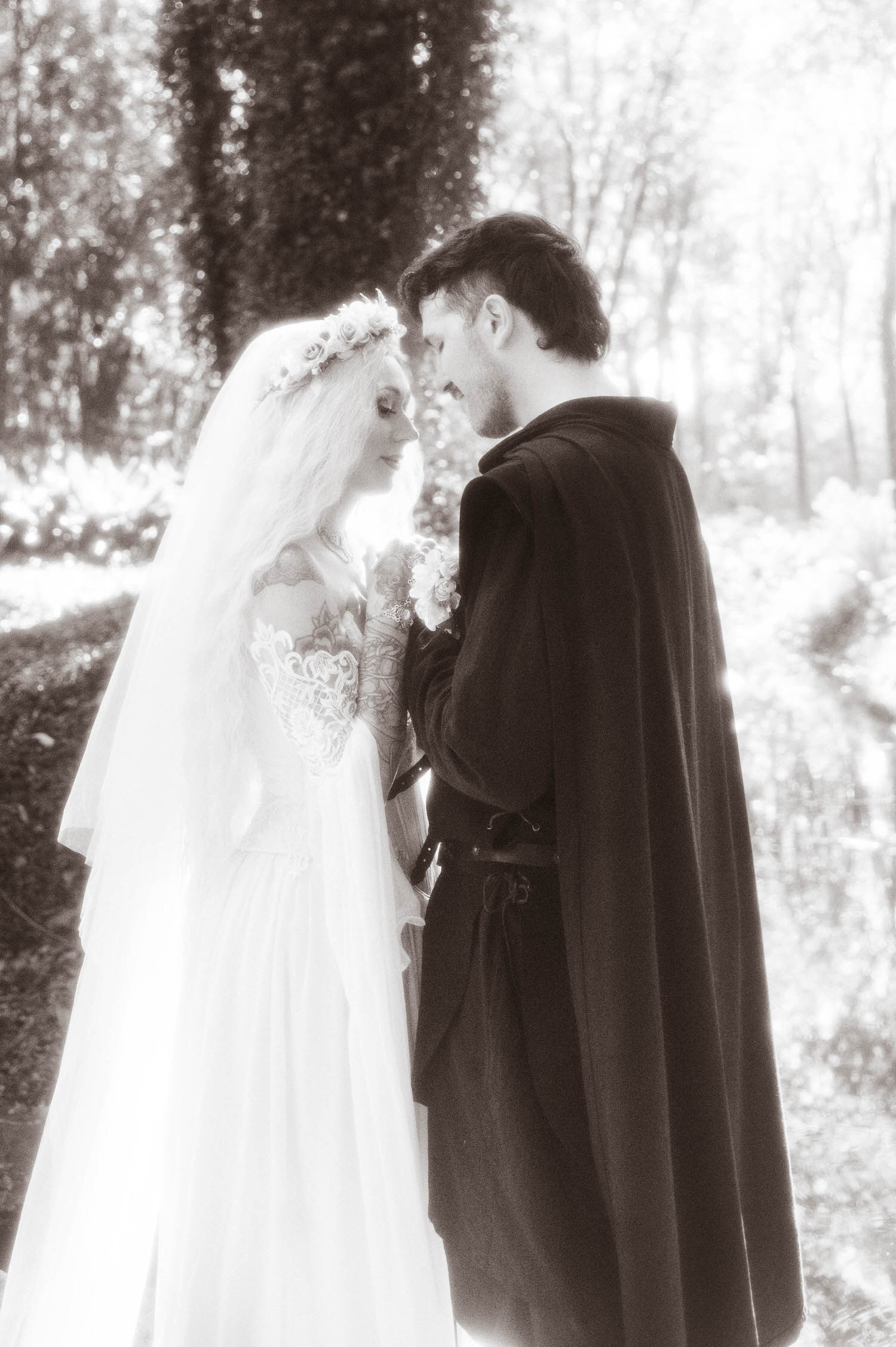 In this black and white portrait, the bride and groom lean close together in the woods, her flower crown and his cloak creating a striking contrast. Classic compositions like this are a signature of a Texas Renaissance Festival wedding photographer. Royal Lune Photo.