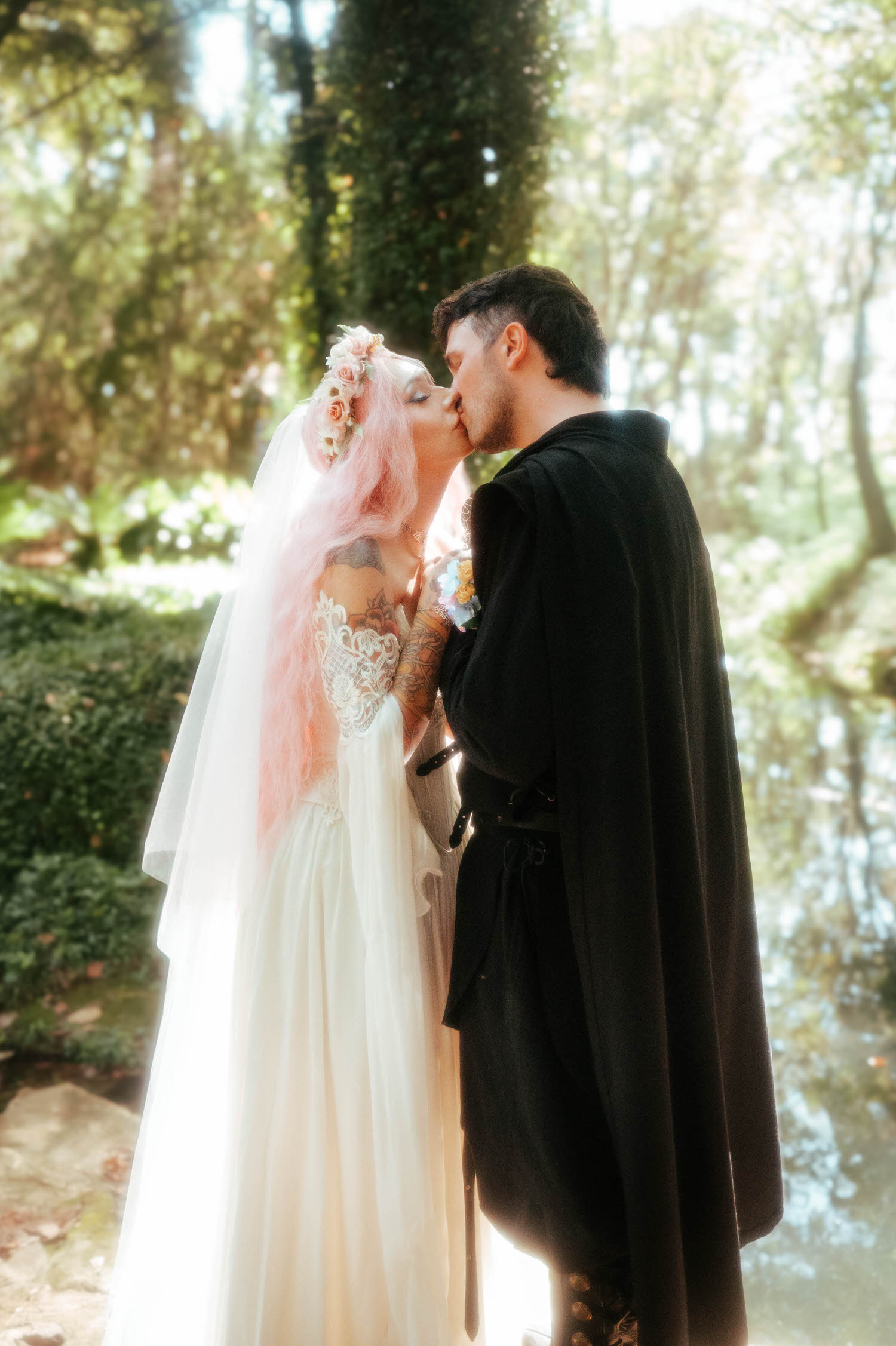The newlyweds share a kiss beside the pond, the bride's flower crown and the groom's black cloak framed by soft greenery and reflections in the water. This peaceful setting is a favorite for a fantasy wedding photographer. Royal Lune Photo.