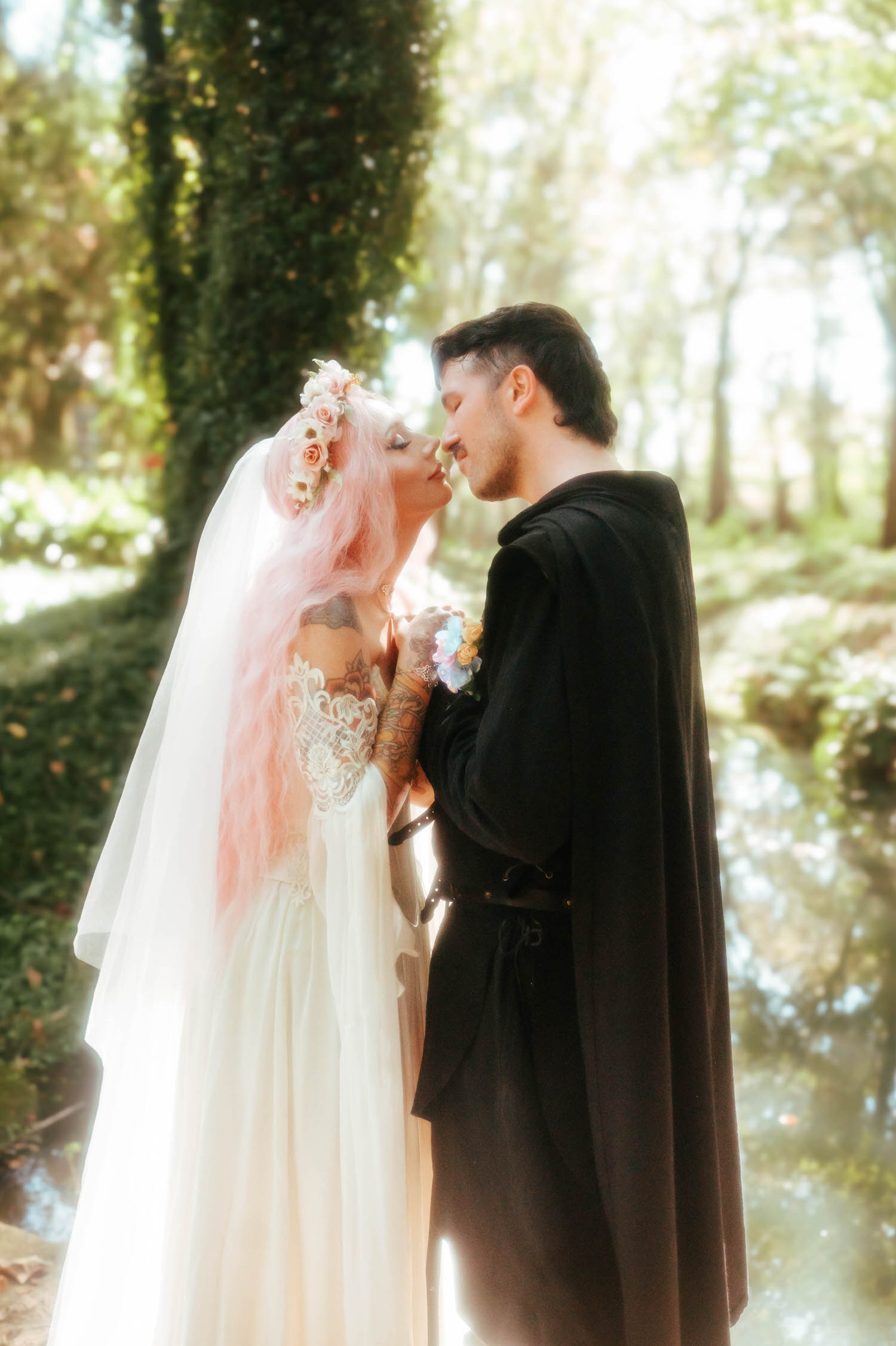 The bride and groom stand nose to nose beside a quiet pond, her pink hair cascading over her shoulder as he holds her gently. Serene, natural backdrops like this make for a dreamy Renaissance festival wedding. Royal Lune Photo.