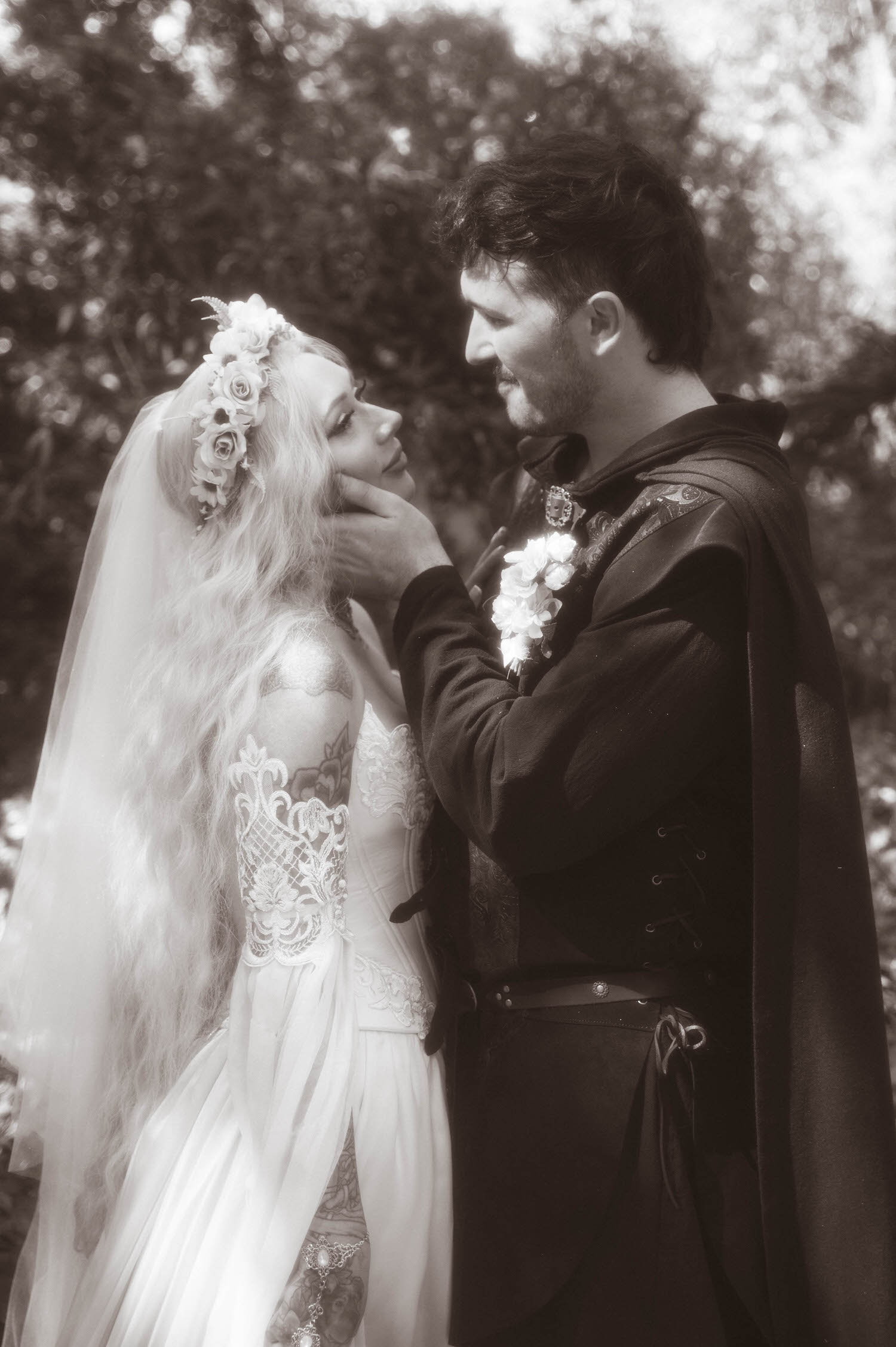 In this black and white portrait, the groom holds the bride's chin as they share an intimate look, her long veil and flower crown flowing in the breeze. Timeless details like this highlight the artistry of a Texas Renaissance Festival wedding photographer. Royal Lune Photo.