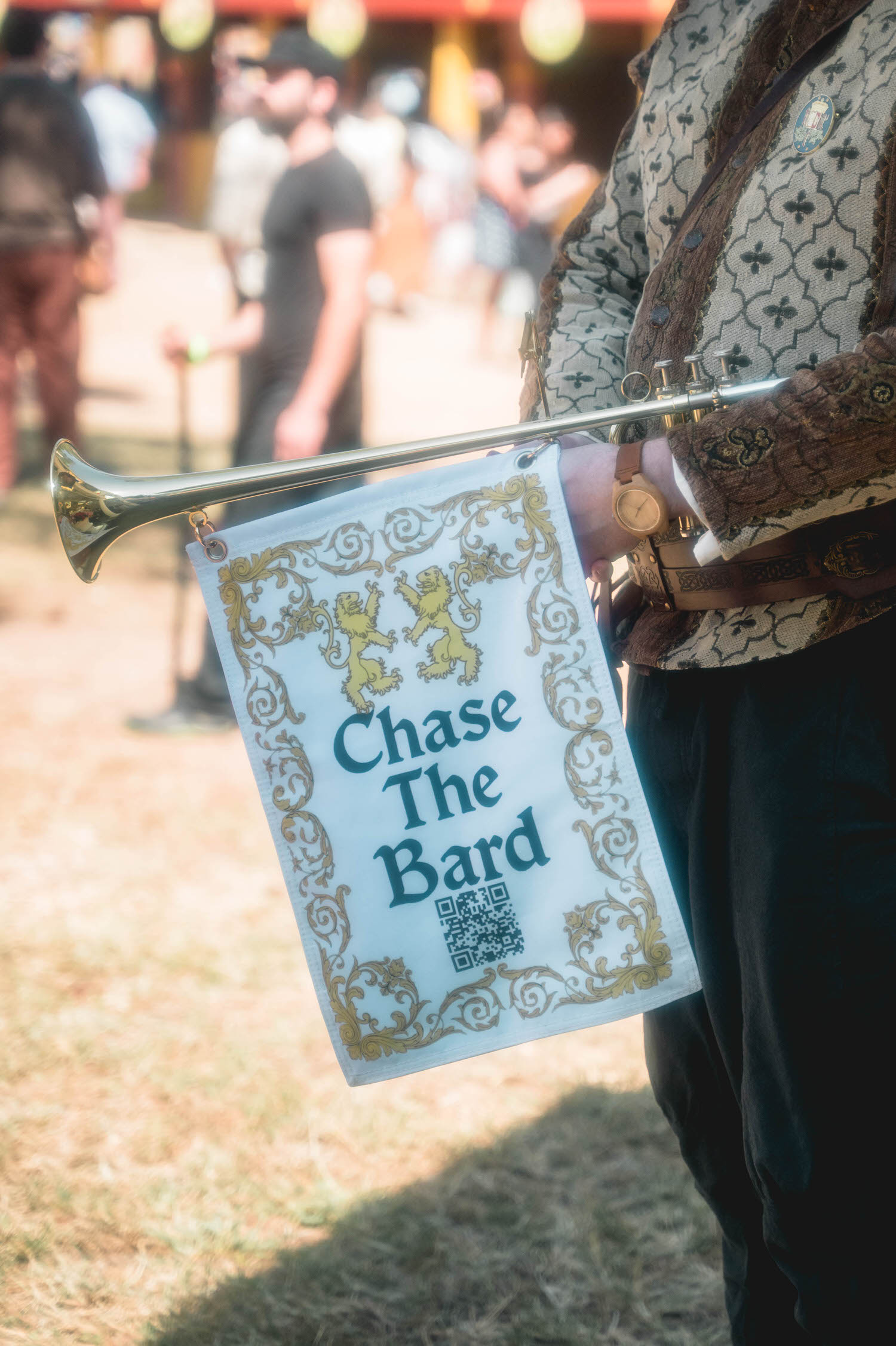 A festival performer carries a trumpet decorated with a small banner reading Chase The Bard, adding playful Renaissance character to the wedding day. Touches like this bring authentic festival charm to a Renaissance festival wedding. Royal Lune Photo.