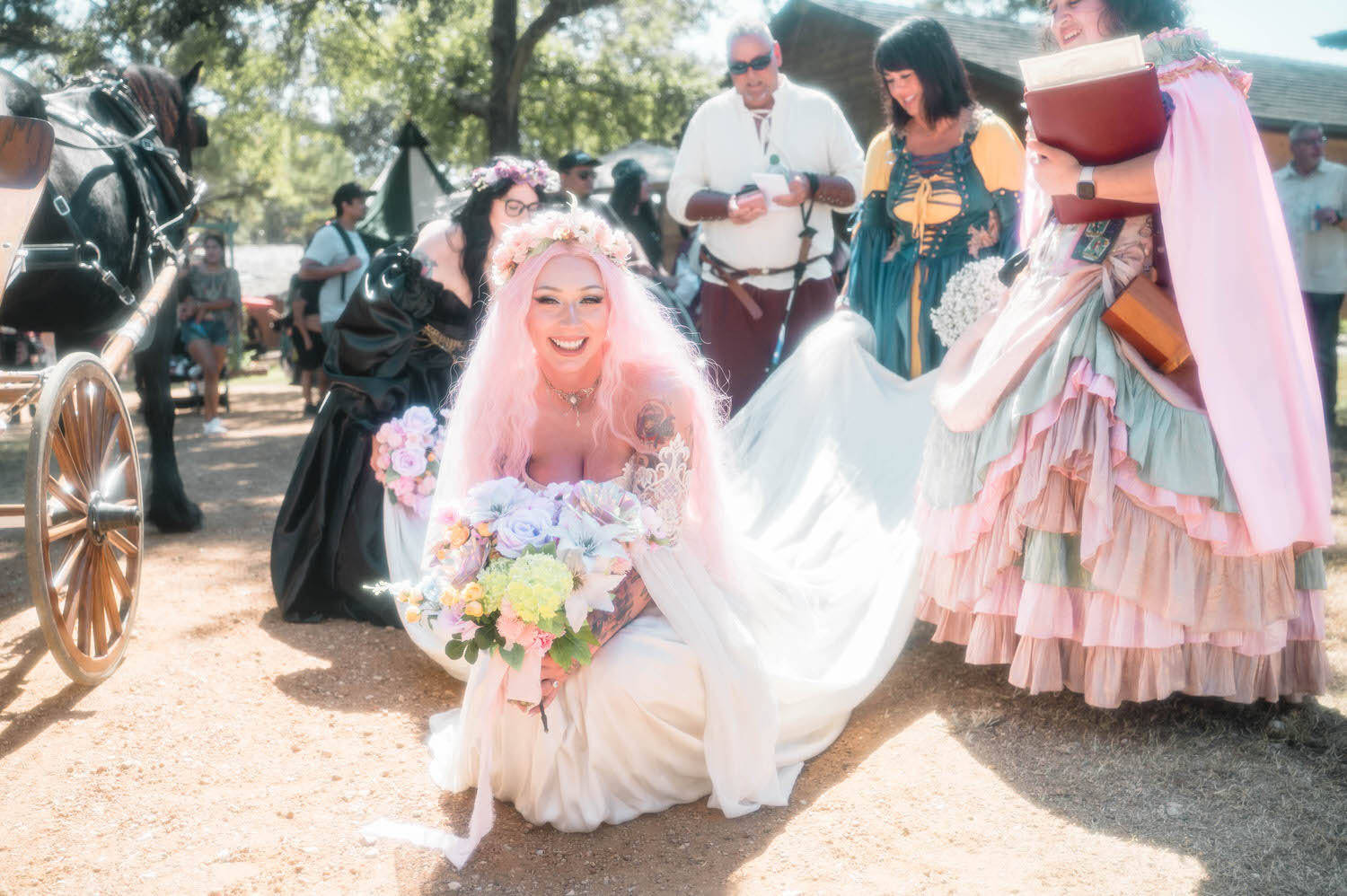 The bride kneels on the ground in her Firefly Path Bridal Gown, laughing with her bridesmaids as her veil and gown spill across the dirt path. The flowing silhouette and lace sleeves make this gown a stunning choice for a Renaissance festival wedding. Royal Lune Photo.