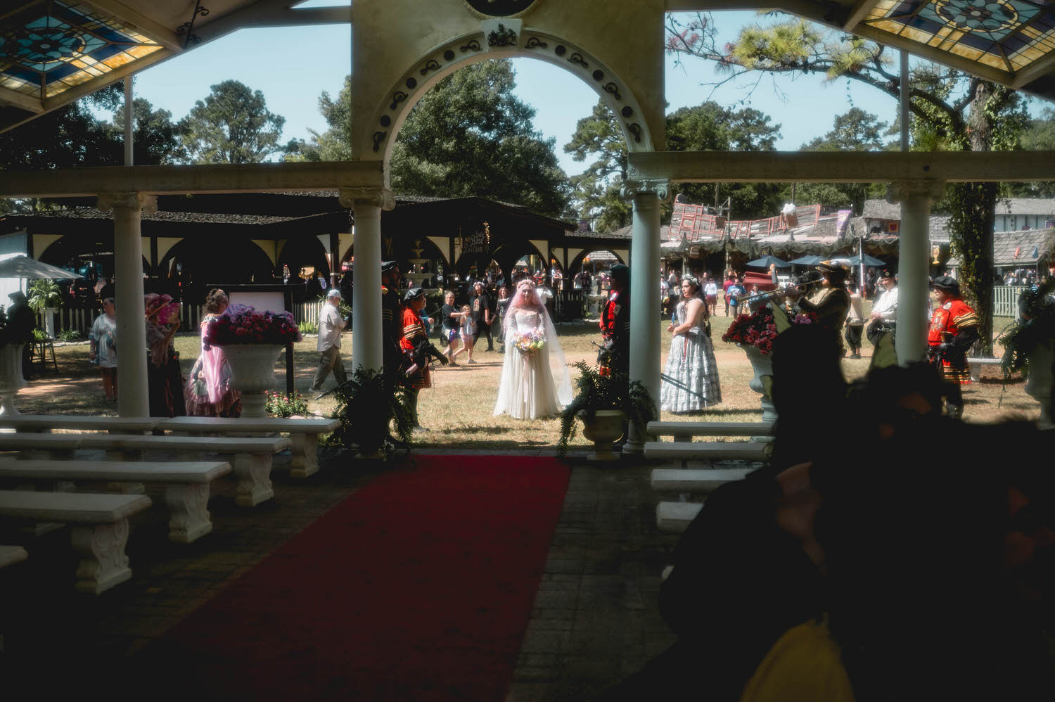 The bride smiles softly while walking the red carpet toward the altar, her flower crown and pink hair glowing in the sunlight as her bouquet catches the light. Moments like this are why couples search for a Texas Renaissance Festival wedding photographer who can capture the romance of the day. Royal Lune Photo.