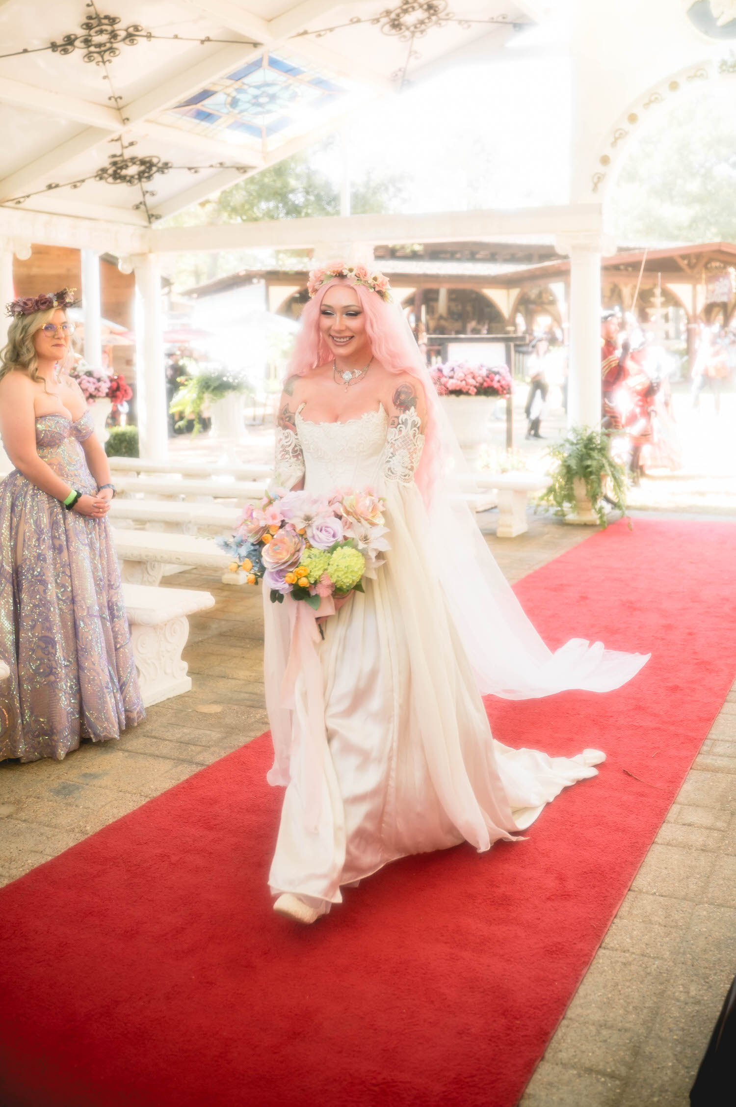 The bride walks the red carpet aisle past white stone benches and stained glass windows, her bridesmaid in an iridescent gown watching from the side. The Basilica Wedding Chapel offers a stunning backdrop for every step of the processional. Royal Lune Photo.