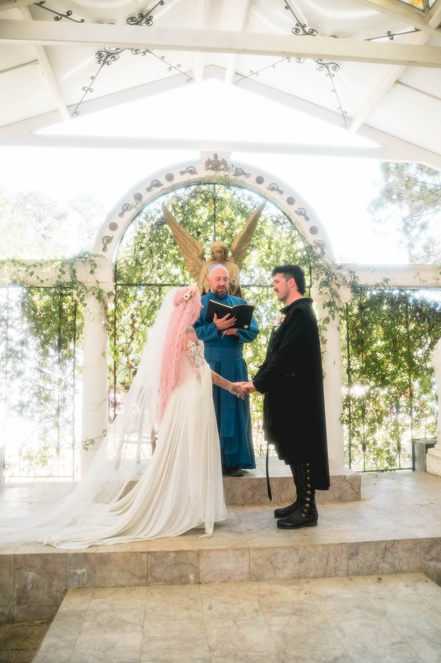 The bride and groom hold hands at the altar beneath a golden angel statue draped in greenery while the officiant in a blue robe reads from his book. This dramatic altar setting is a favorite for couples planning a Renaissance festival wedding. Royal Lune Photo.