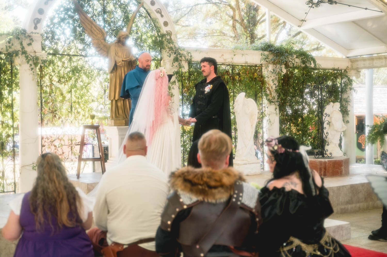 Guests in elaborate Renaissance costumes watch the ceremony from the front row as the bride and groom exchange vows beneath the golden angel statue. The mix of fantasy attire and natural greenery makes this a dream setting for a fantasy wedding photographer. Royal Lune Photo.