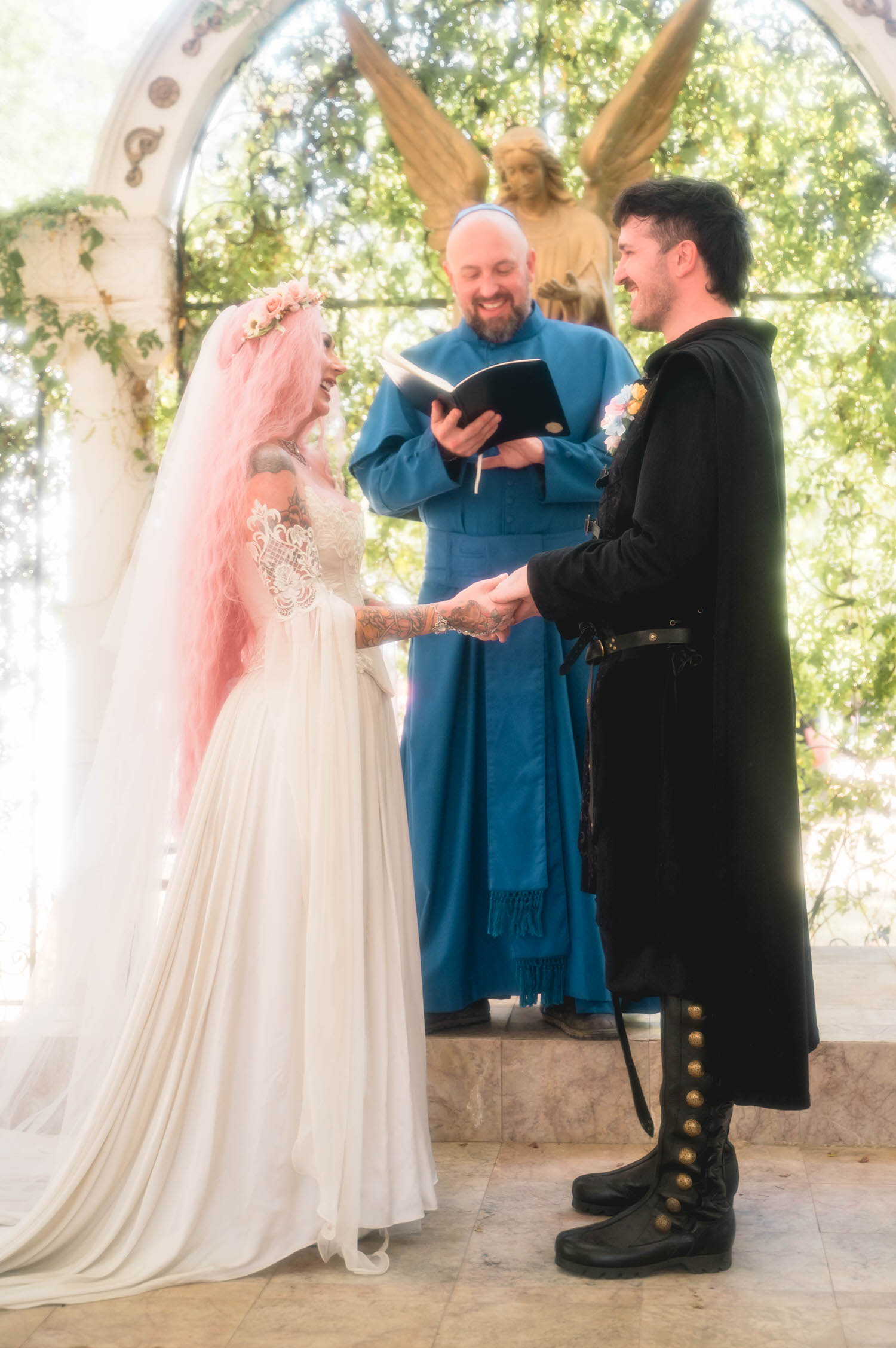 The bride and groom share a laugh while holding hands at the altar, the officiant smiling behind them as the golden angel statue rises above the ivy covered archway. Genuine, joyful moments like this define a Texas Renaissance Festival wedding. Royal Lune Photo.