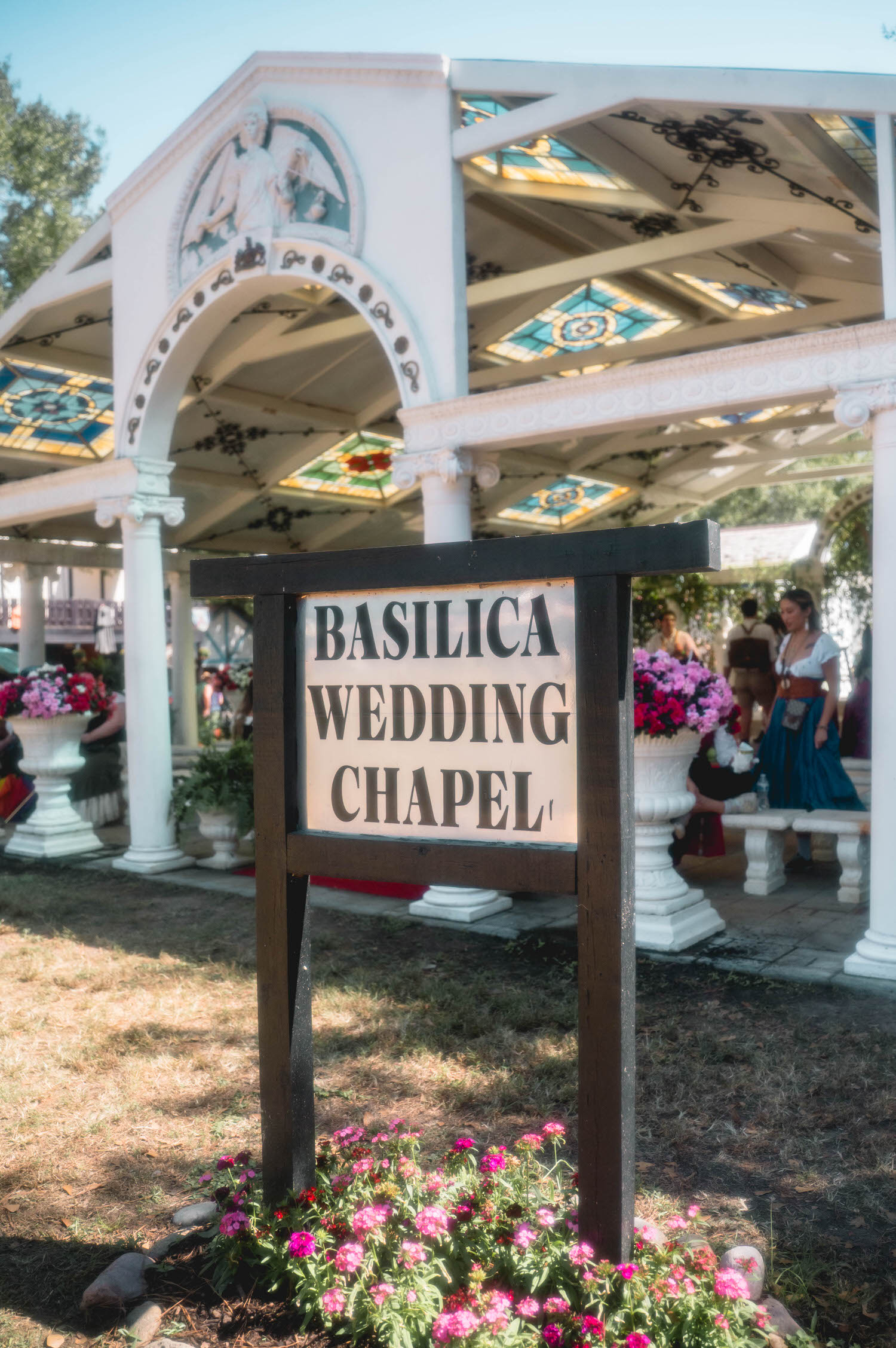 The Basilica Wedding Chapel sign stands among pink flowers in front of the ornate stained glass pavilion at the Texas Renaissance Festival. This iconic ceremony venue is a favorite backdrop for couples planning a Texas Renaissance Festival wedding. Royal Lune Photo.