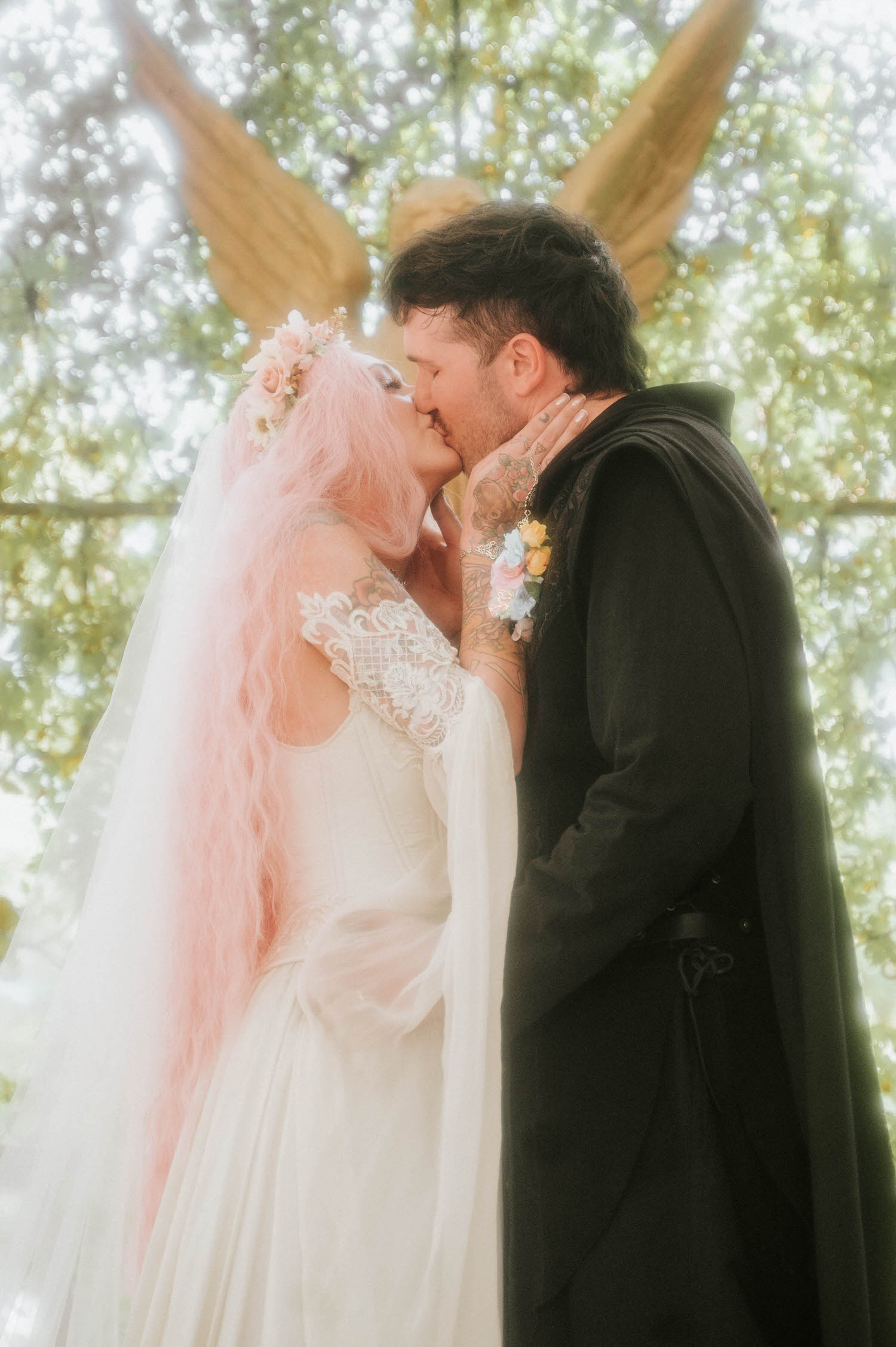 The newlyweds kiss beneath the arched chapel entrance, the bride's pink hair and flowing veil contrasting beautifully with the groom's black cloak and gold buttoned boots. This striking color palette is a signature look for a Renaissance festival wedding. Royal Lune Photo.