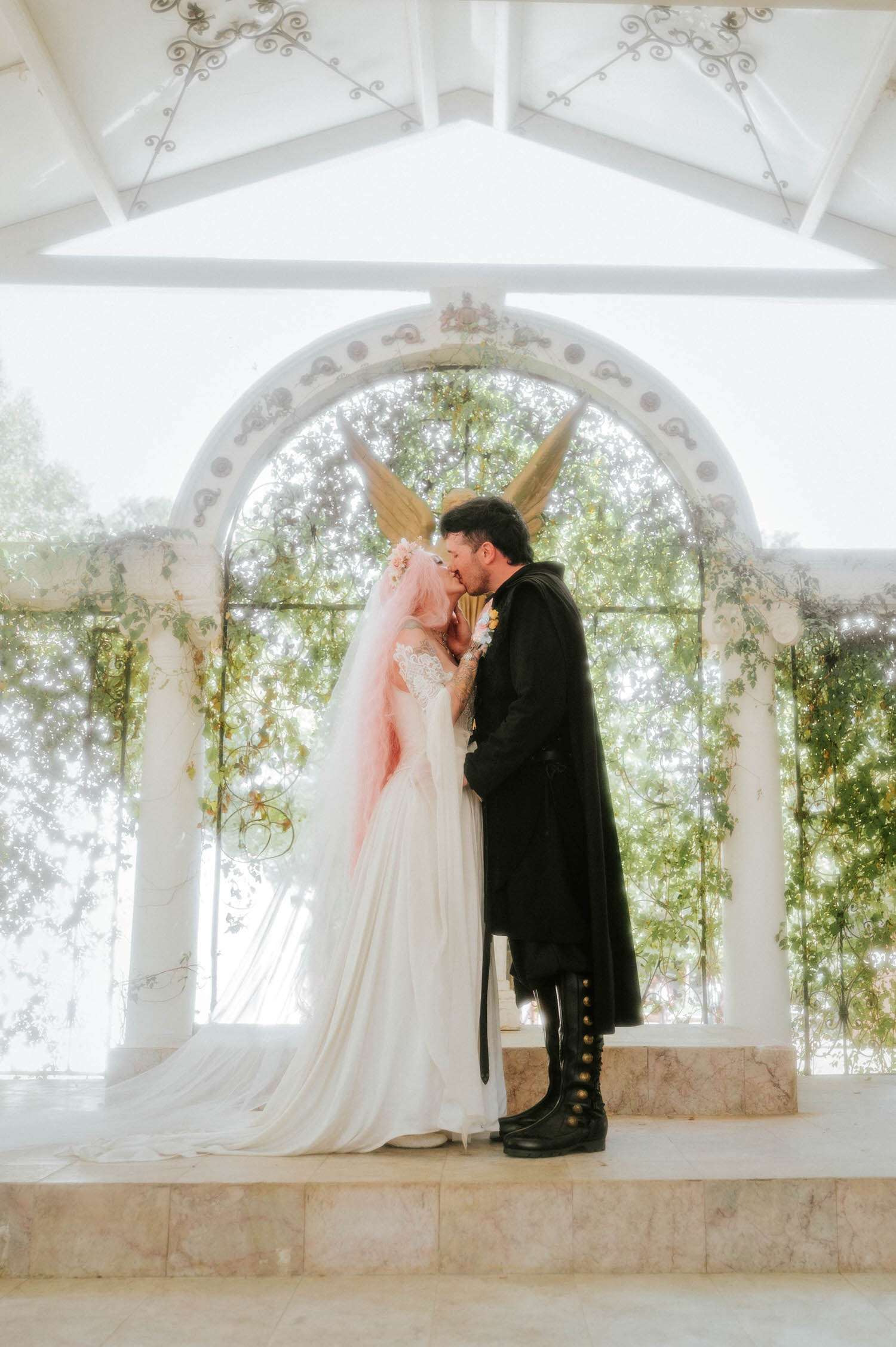 The bride and groom share their first kiss as husband and wife, framed by the golden angel statue and lush greenery climbing the chapel archway. This is the kind of unforgettable moment couples hope for from their Texas Renaissance Festival wedding photographer. Royal Lune Photo.