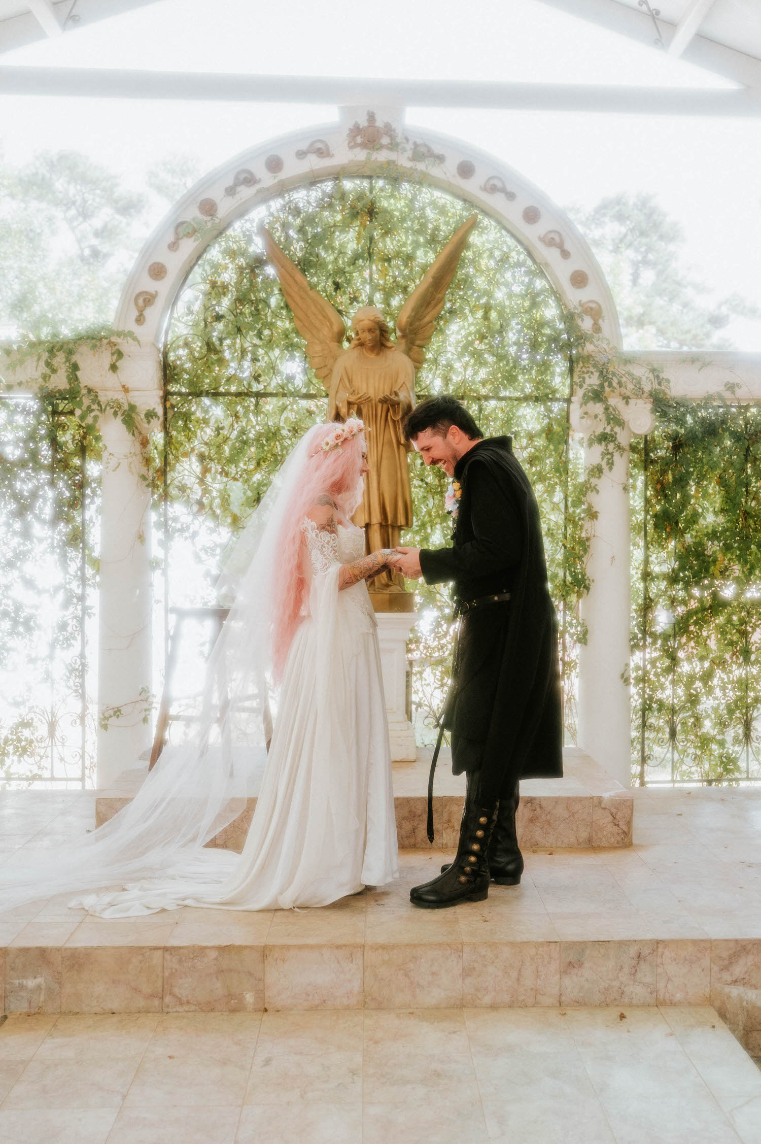 The groom slides the wedding ring onto the bride's finger as they smile at each other beneath the golden angel statue at the altar. Intimate details like this are an essential part of documenting a Texas Renaissance Festival wedding. Royal Lune Photo.