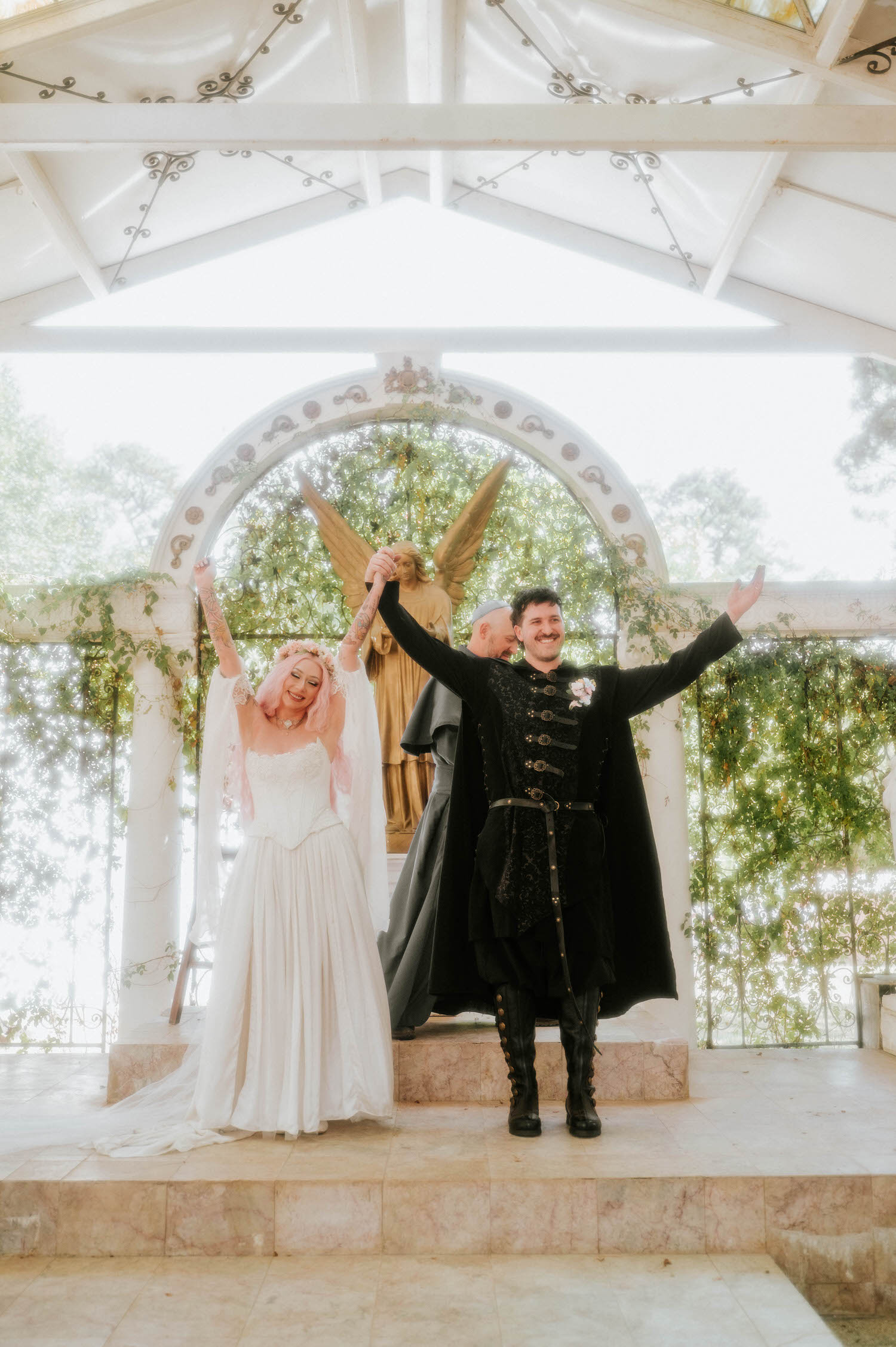 The bride and groom raise their joined hands in celebration at the altar as the officiant smiles behind them, the golden angel statue framed by greenery overhead. Triumphant moments like this are a highlight of any Texas Renaissance Festival wedding. Royal Lune Photo.