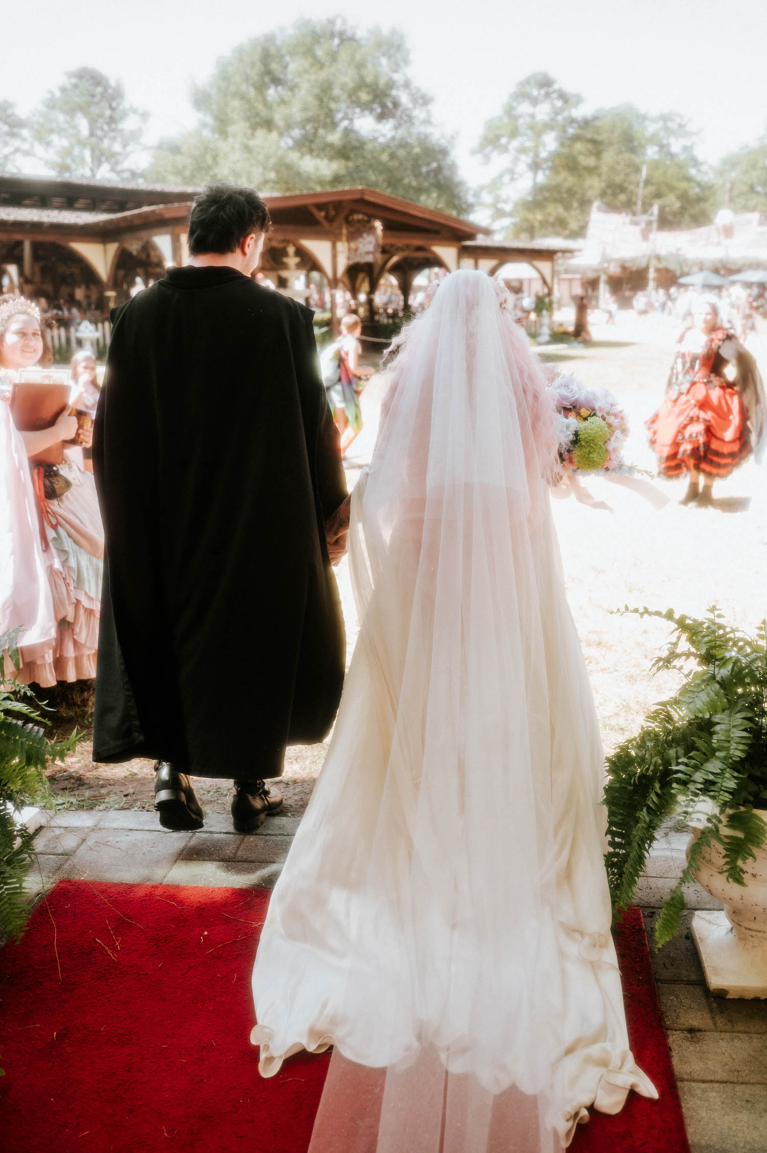 The bride and groom walk hand in hand away from the chapel, her veil and gown trailing across the red carpet as festival guests look on in the background. Quiet moments like this after the ceremony are part of what makes a Texas Renaissance Festival wedding photographer's storytelling so complete. Royal Lune Photo.
