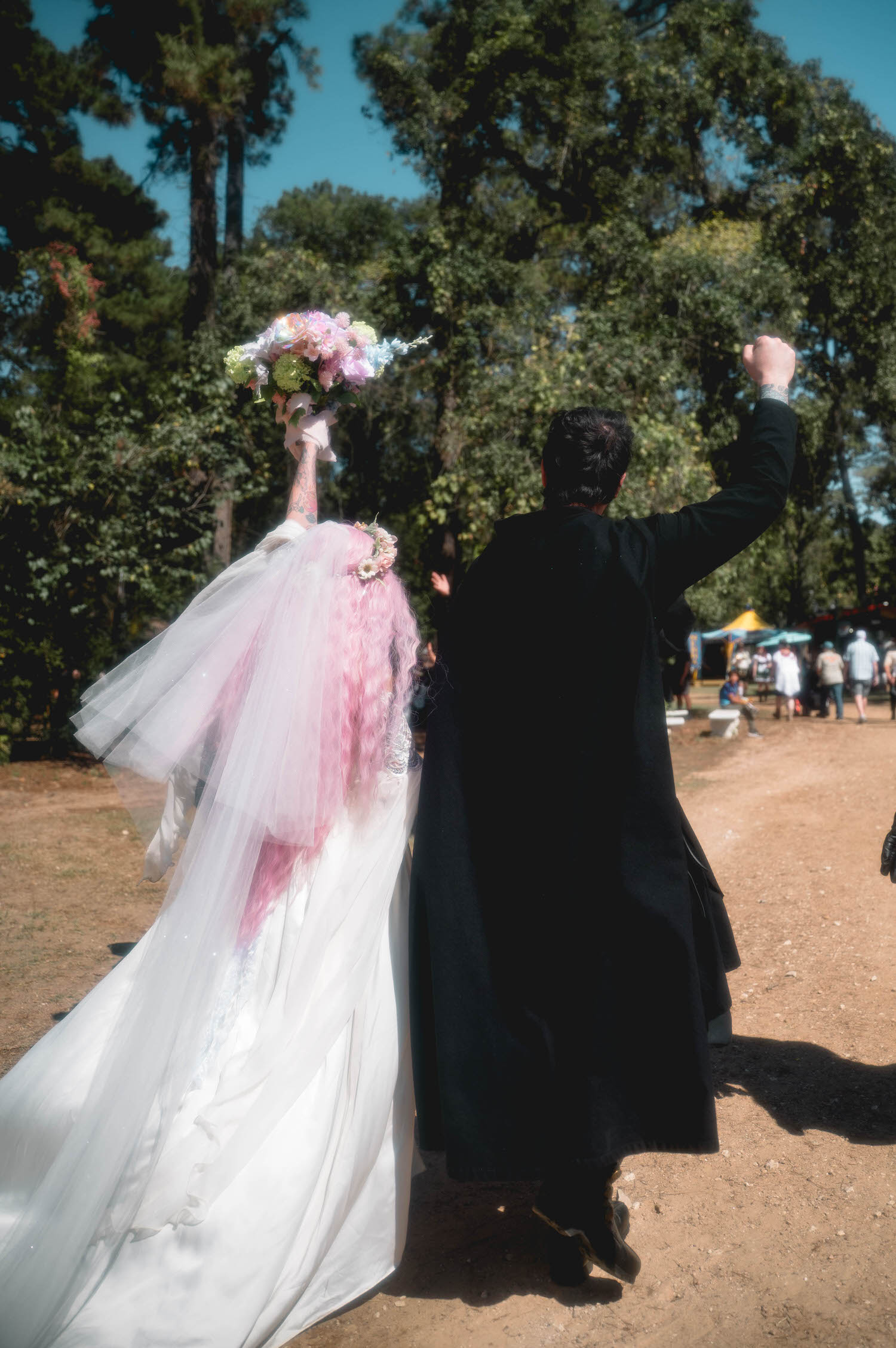 The bride raises her bouquet high while the groom lifts his fist in triumph, both walking away from the camera through the festival grounds. Joyful, high energy moments like this capture the true spirit of a Texas Renaissance Festival wedding. Royal Lune Photo.