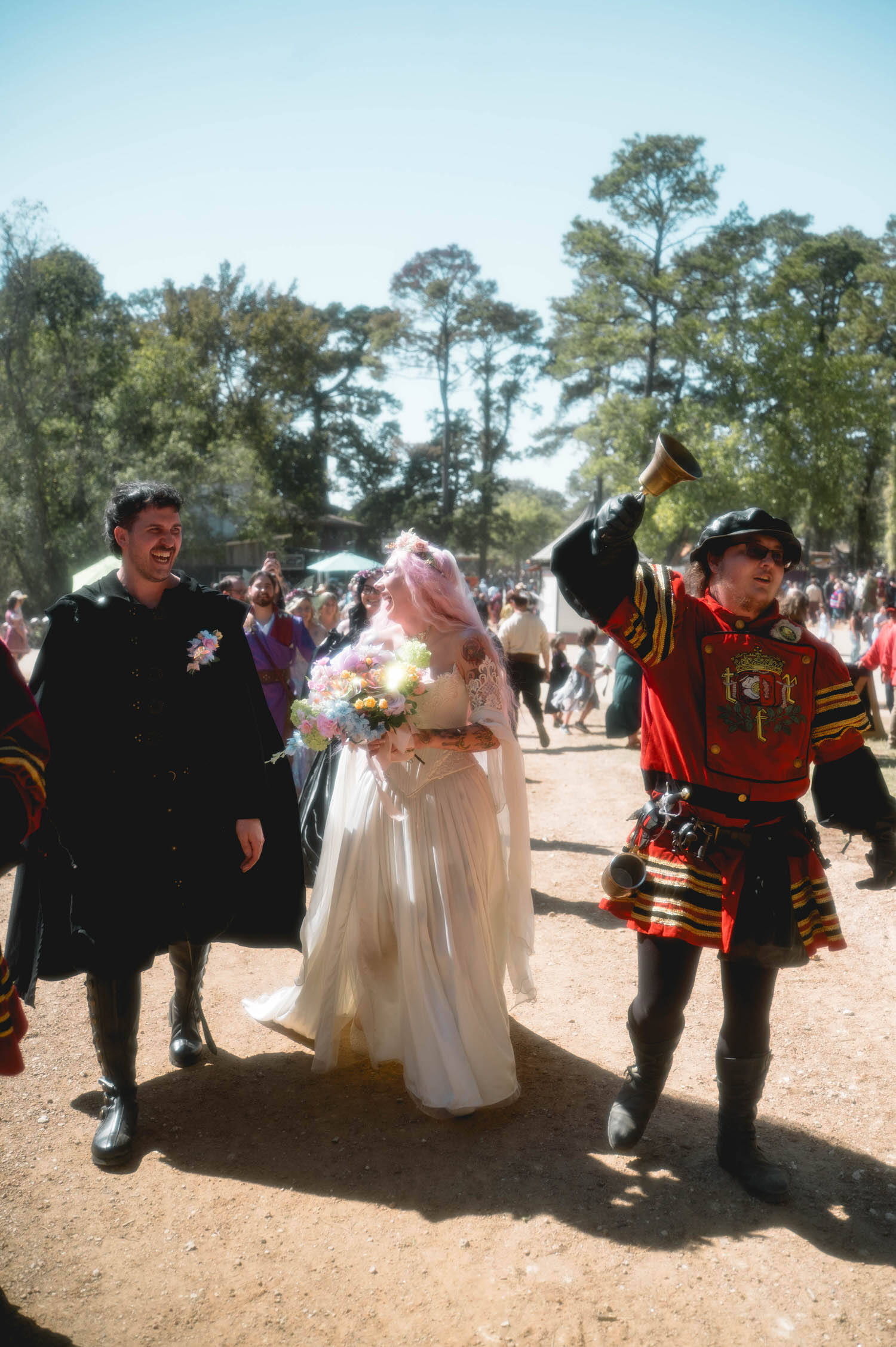 The newlyweds walk together laughing as a festival town crier rings a bell beside them, announcing their celebration to the crowd. Playful interactions like this are a hallmark of a Renaissance festival wedding. Royal Lune Photo.
