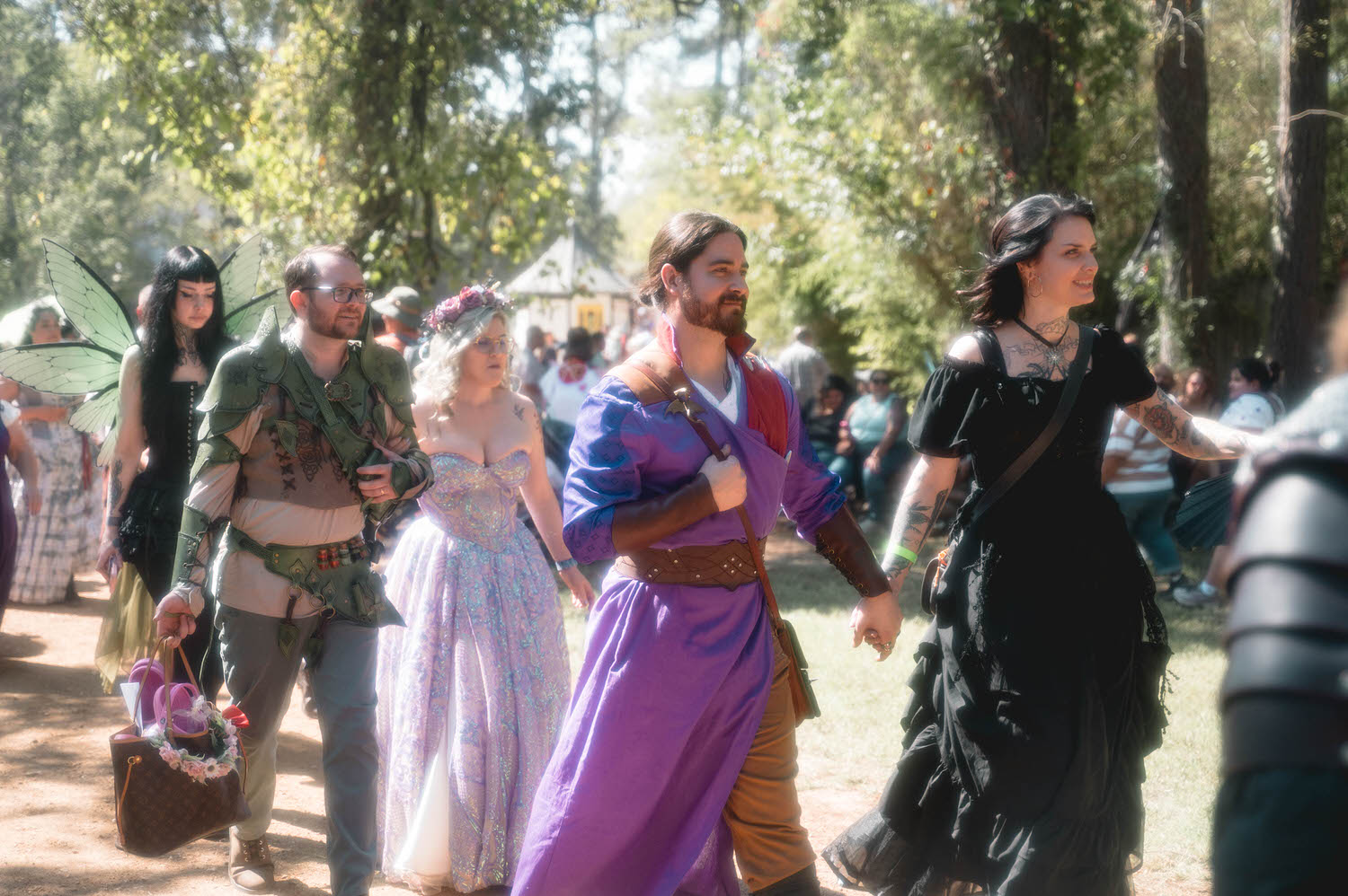 A colorful group of festival guests in fairy wings, medieval gowns, and Renaissance attire walk together through the wooded festival grounds. Scenes like this showcase the vibrant community that makes a Texas Renaissance Festival wedding so unique. Royal Lune Photo.
