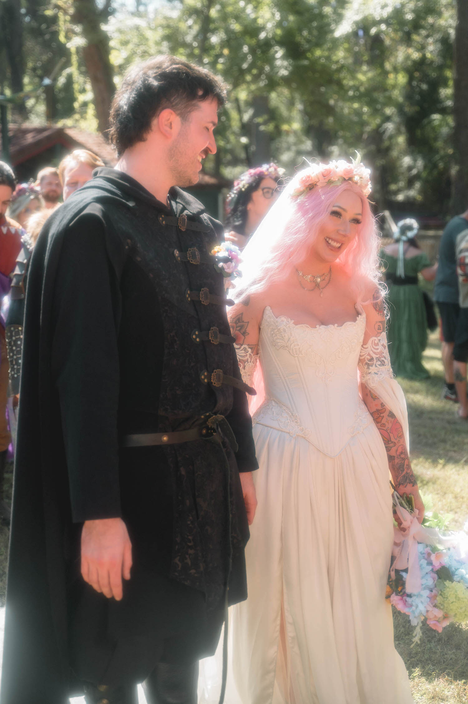 The groom looks at the bride with a warm smile as she laughs, her lace sleeves and tattooed arms catching the afternoon light among the festival crowd. Genuine moments like this are a hallmark of a skilled Texas Renaissance Festival wedding photographer. Royal Lune Photo.