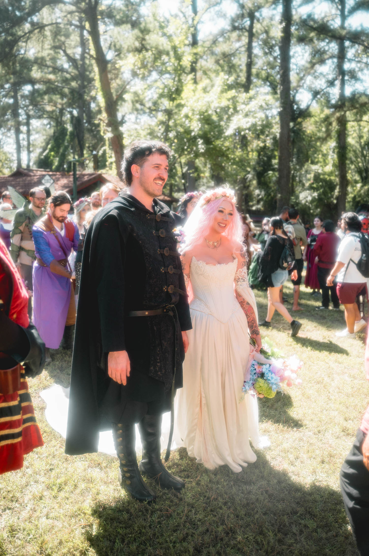 The groom smiles beside the bride as festival guests in medieval costume mingle around them, her pink hair and floral crown glowing in the sunlight. Candid crowd shots like this capture the festive energy of a Texas Renaissance Festival wedding. Royal Lune Photo.