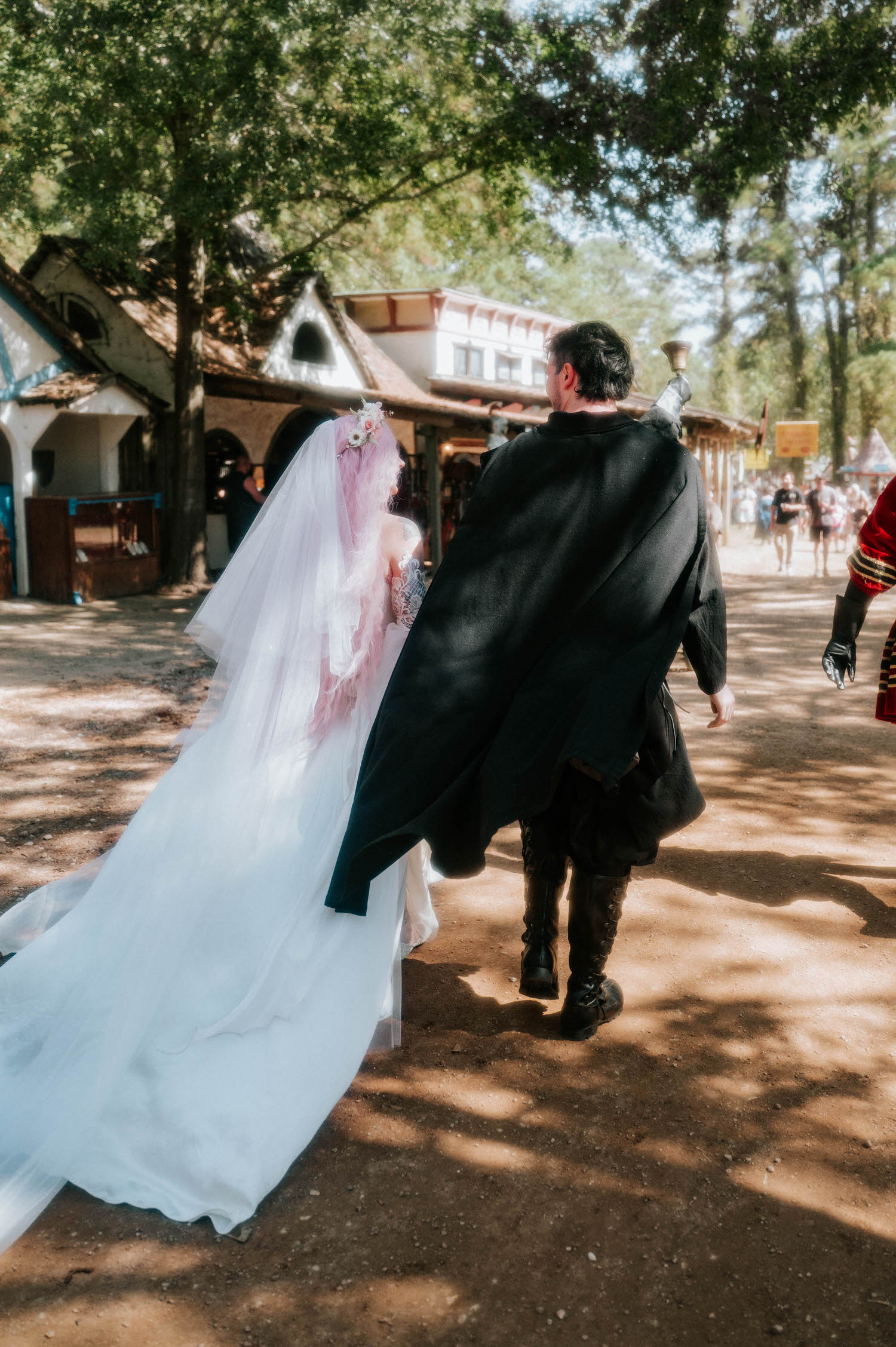 The newlyweds walk arm in arm past festival shops, the bride's veil and gown trailing across the dirt path as festival goers look on. Candid moments like this capture the lively atmosphere of a Texas Renaissance Festival wedding. Royal Lune Photo.