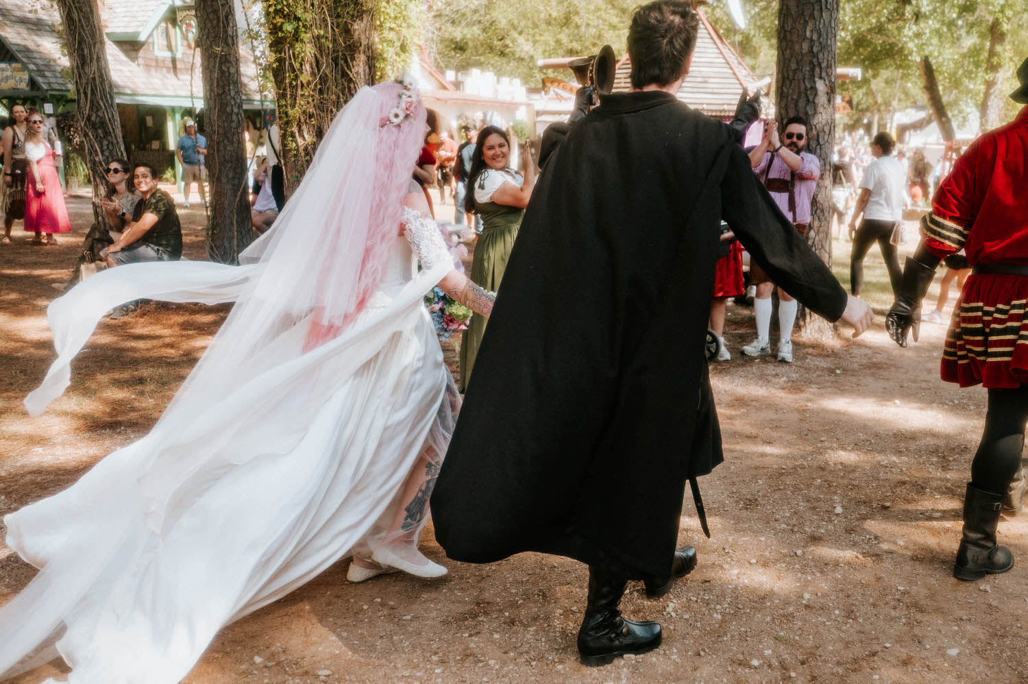 The bride's veil billows in the wind as the groom raises a trumpet in celebration, festival guests clapping and cheering around them. Spontaneous moments like this are part of what makes a Texas Renaissance Festival wedding photographer's job so fun. Royal Lune Photo.