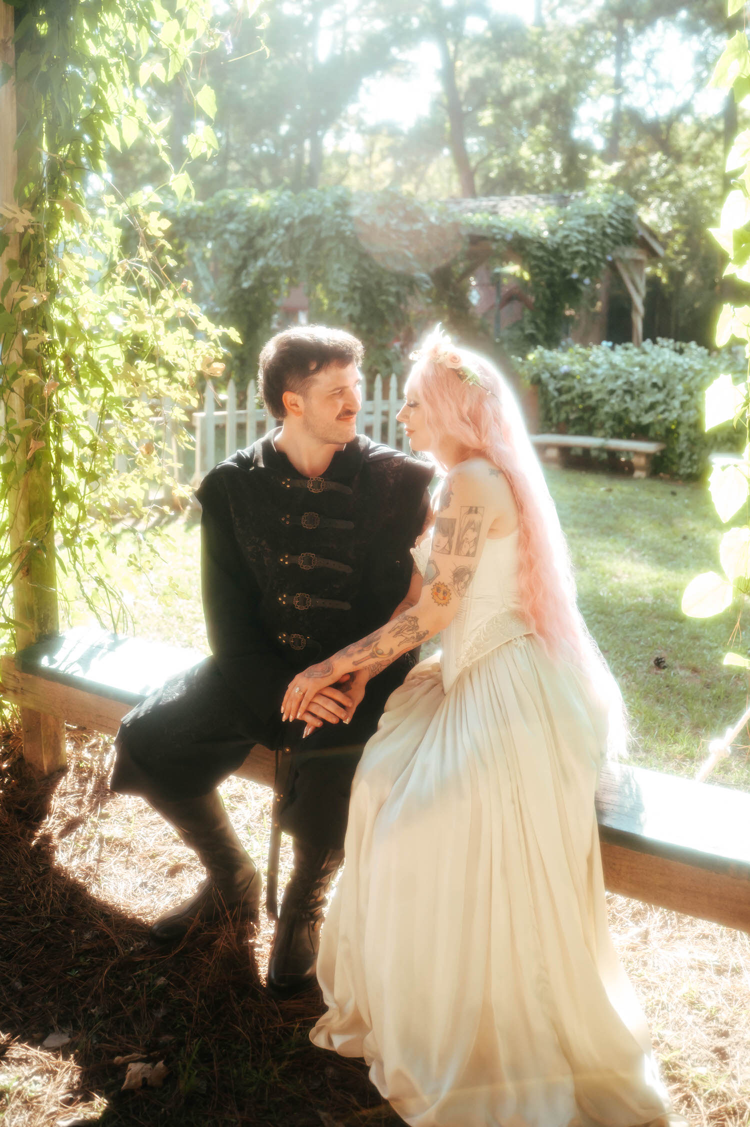 The bride and groom sit close together on a vine covered bench, her pink hair and tattooed arm resting against his black medieval coat as they gaze at each other. Quiet, intimate moments like this round out the story of a Texas Renaissance Festival wedding. Royal Lune Photo.