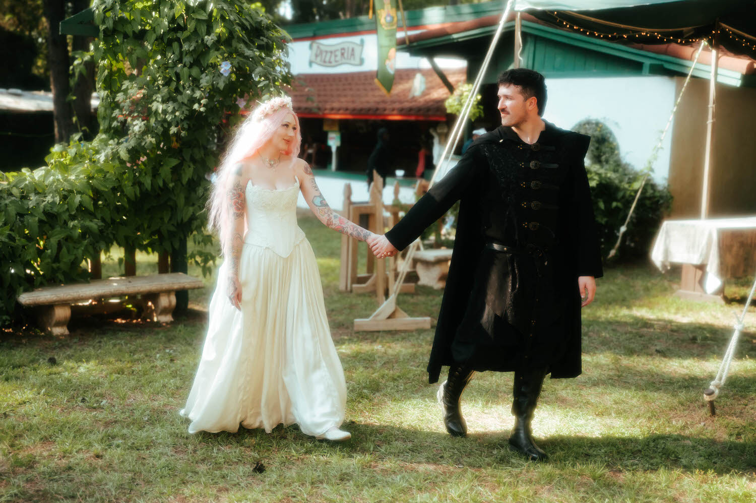 The newlyweds walk hand in hand past a pizzeria booth and reception tent, her ivory gown trailing across the grass. Candid in between moments like this capture the full atmosphere of a Texas Renaissance Festival wedding. Royal Lune Photo.