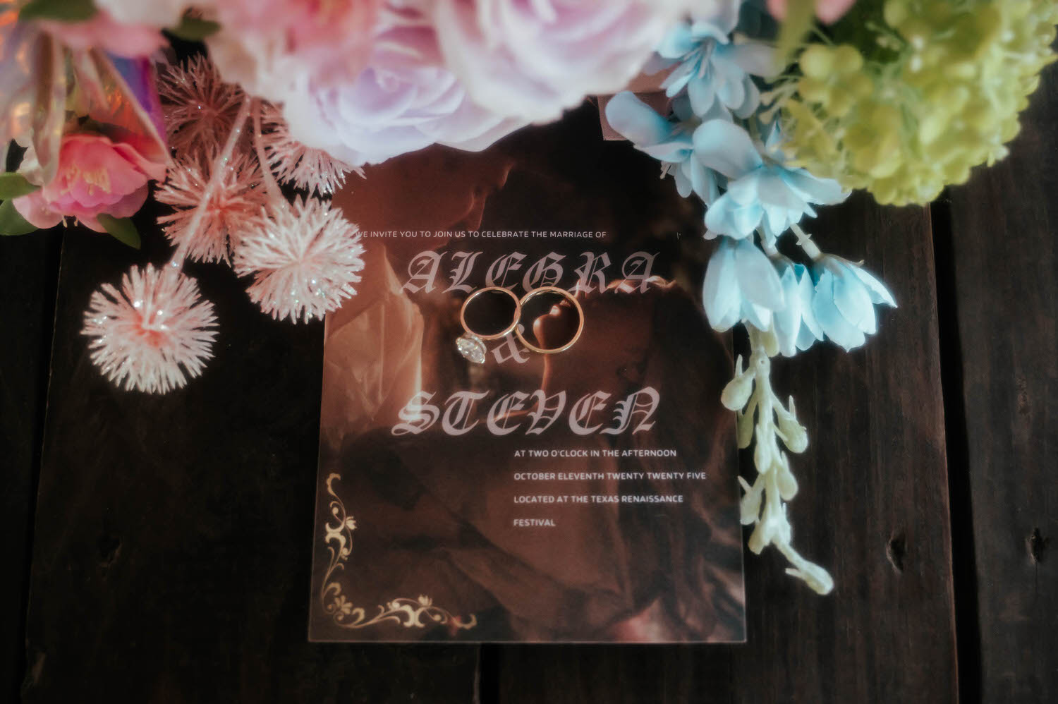 The wedding invitation for Alegra and Steven rests beneath floral arrangements with gold wedding bands tucked into the design, noting the celebration at the Texas Renaissance Festival. Stationery details like this help tell the full story of a Texas Renaissance Festival wedding. Royal Lune Photo.
