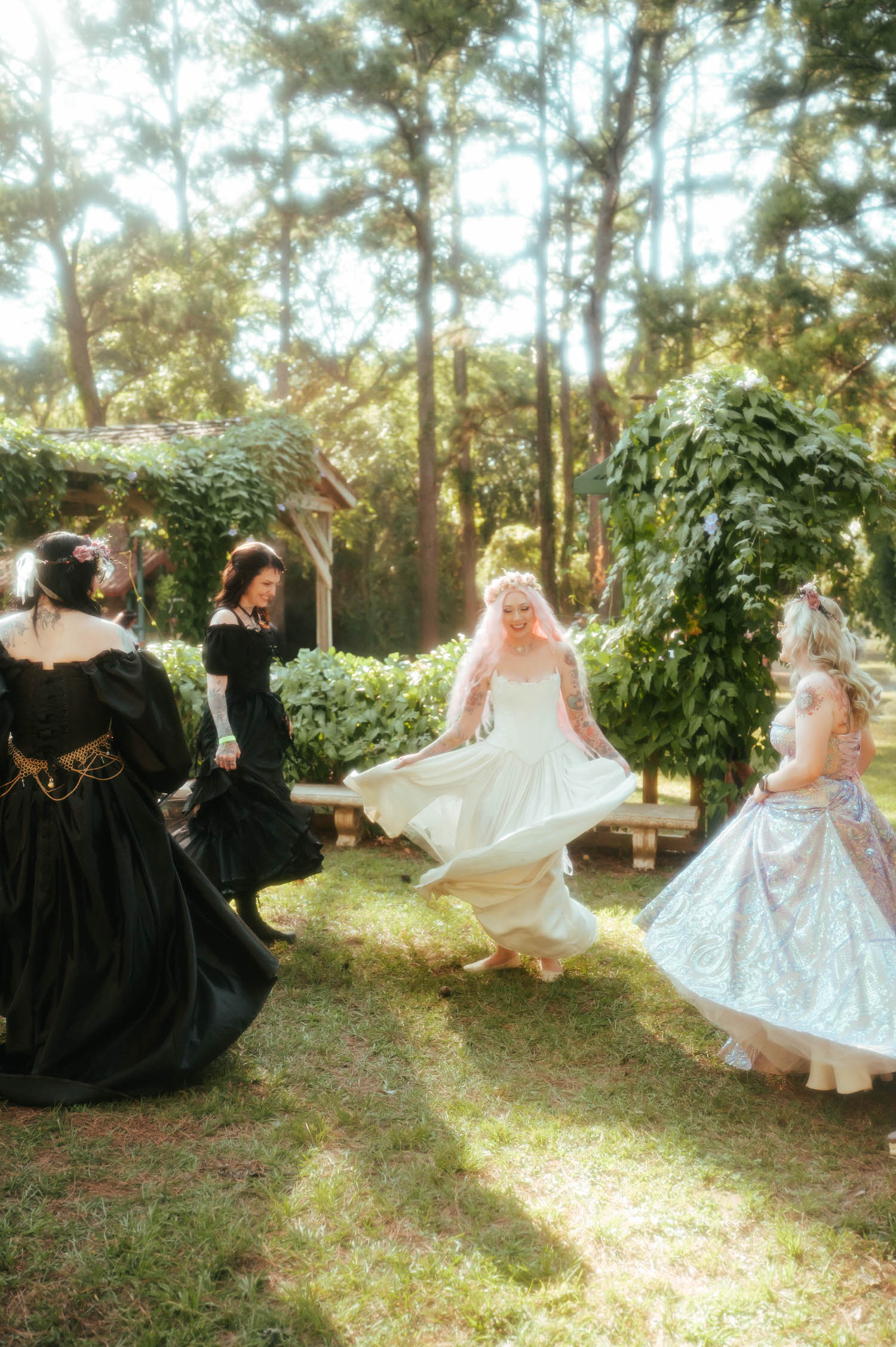 The bride laughs with her bridal party near a stone bench, her pink hair and flower crown glowing in dappled afternoon light. Royal Lune Photo.