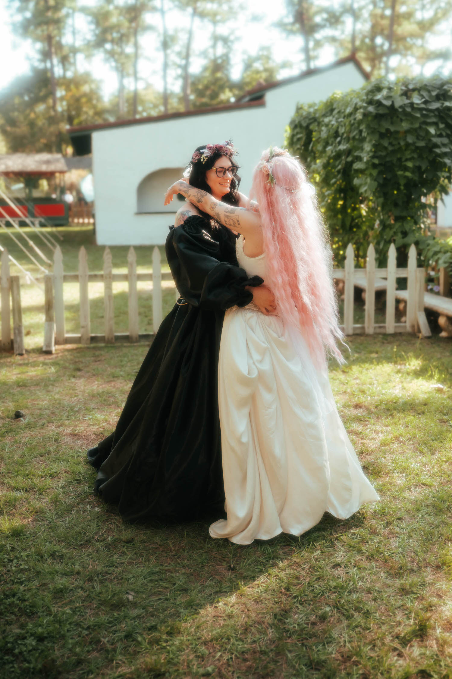 The bride and maid of honor in a black gown share a warm embrace on the lawn, her pink hair flowing against the guest's dark dress. Joyful friendship moments like this are part of the full story of a Texas Renaissance Festival wedding. Royal Lune Photo.