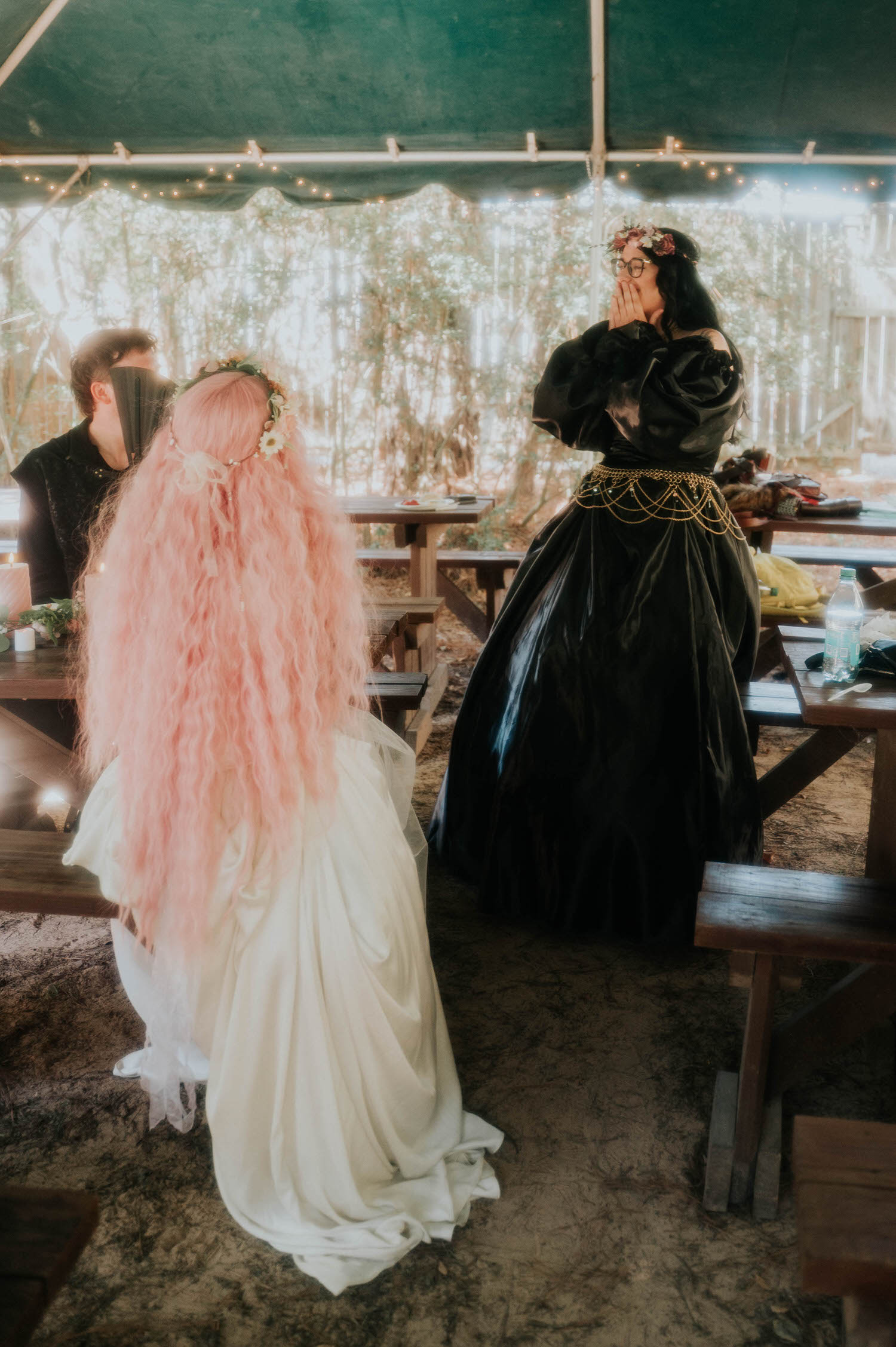The bride's pink hair flows down her back as a guest in a black gown reacts with surprise and joy under the reception tent. Candid emotional moments like this are part of what makes a Texas Renaissance Festival wedding so memorable. Royal Lune Photo.