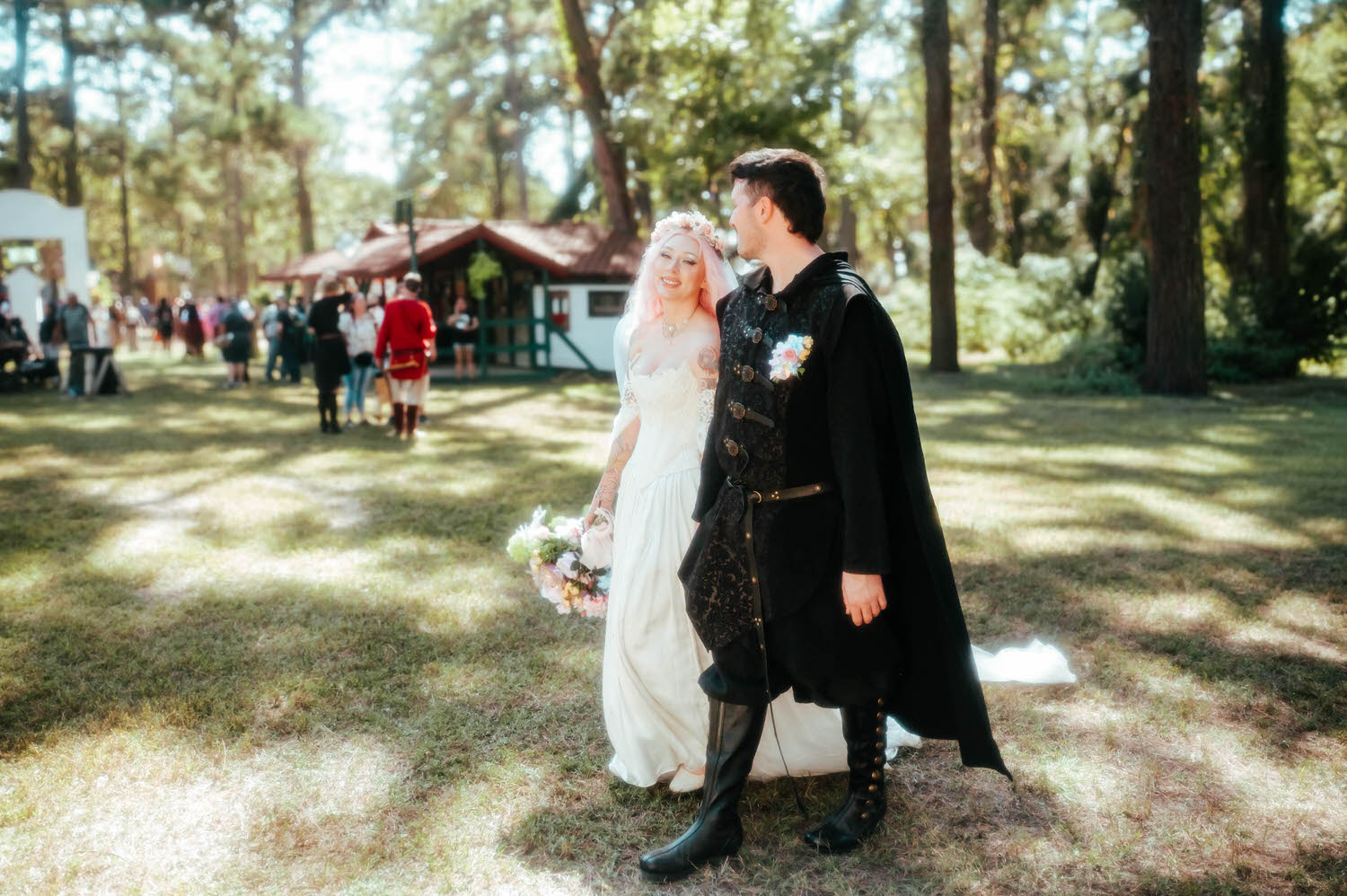 The newlyweds walk hand in hand through a sunlit grove of trees on their way to the reception, festival guests visible in the background. Scenes like this show off the full experience a Texas Renaissance Festival wedding photographer can capture. Royal Lune Photo.