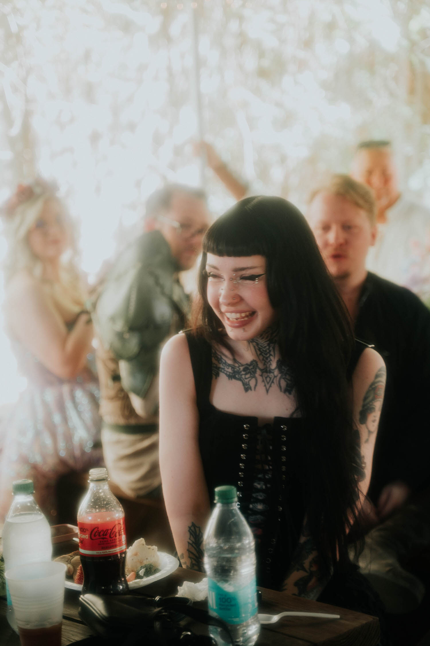 A guest with dark hair and bold tattoos laughs at the reception table, surrounded by drinks and plates of food under the tent. Candid guest moments like this add life to the story of a Texas Renaissance Festival wedding. Royal Lune Photo.