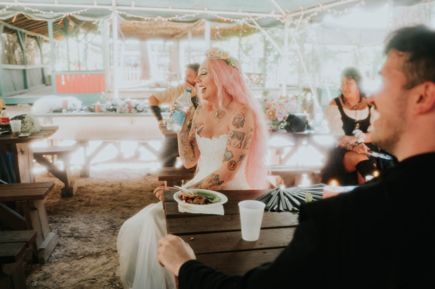The bride laughs while holding a water bottle and enjoying her meal at the reception table, her pink hair and tattoos catching the soft light. Relaxed, joyful moments like this are part of what makes an Italian Village reception so special. Royal Lune Photo.
