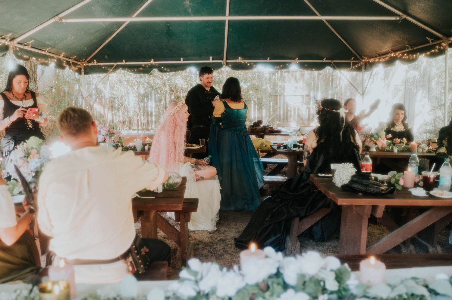 Guests gather under a string lit tent at the Italian Village reception, chatting and laughing around rustic wooden picnic tables decorated with candles and florals. Cozy details like this make the Italian Village reception feel intimate and warm. Royal Lune Photo.