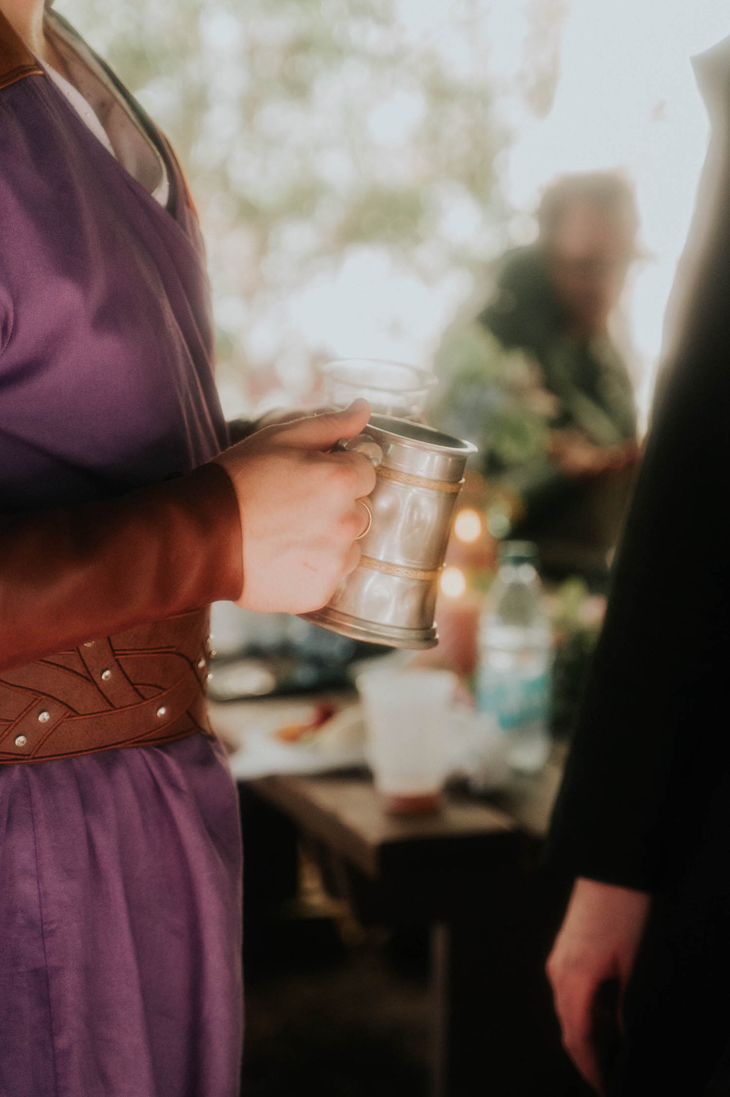 A guest in a purple gown holds a pewter tankard at the reception, festival decor and string lights softly blurred in the background. Small details like this help tell the full story of an Italian Village reception. Royal Lune Photo.
