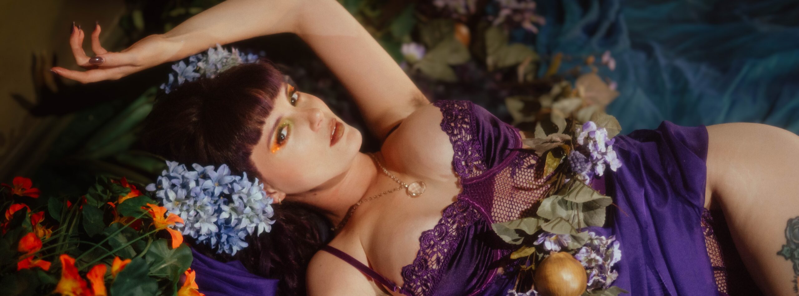 A model reclines among vibrant blue hydrangeas and orange marigolds during a moody boudoir photoshoot, wearing a purple lace and mesh lingerie set with gold and green eyeshadow inspired by the whimsigoth aesthetic, styled at Royal Lune Photo.