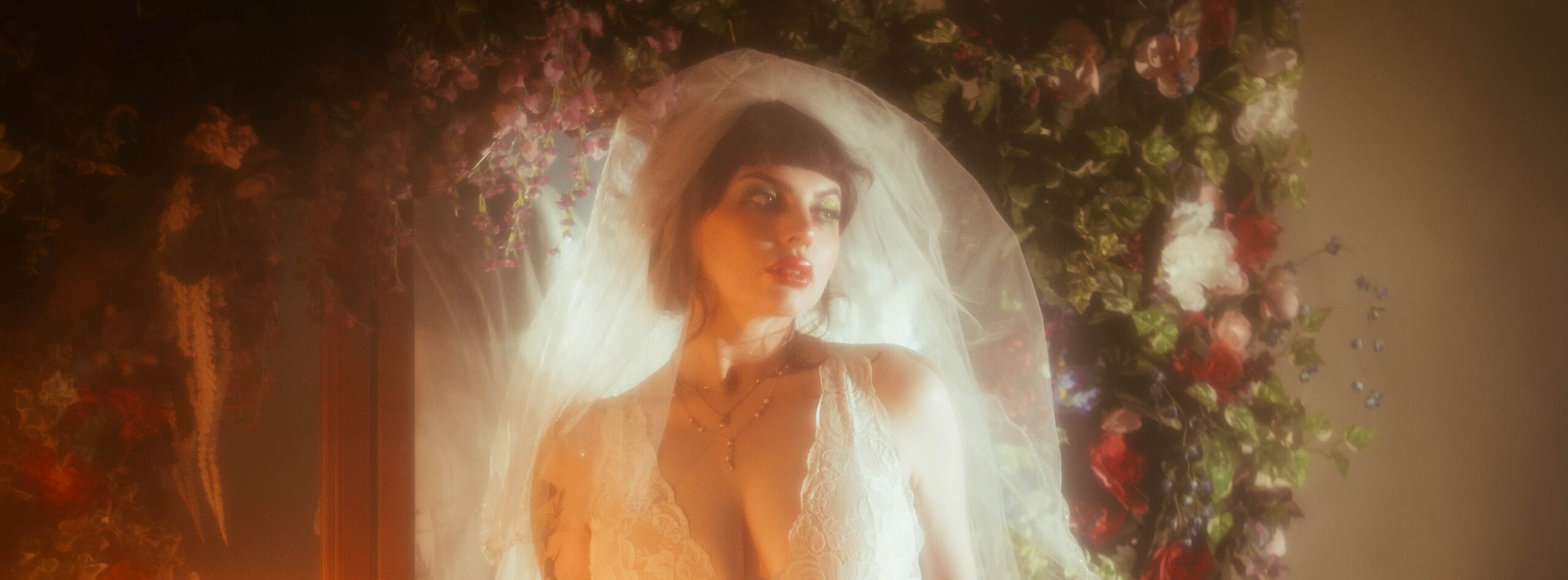 A bride wearing a vintage nightgown boudoir look with a full tulle veil stands in warm, hazy fog machine boudoir photography light in front of a wall of red and white florals, her green eyeshadow and soft glam catching the golden glow at Royal Lune Photo.